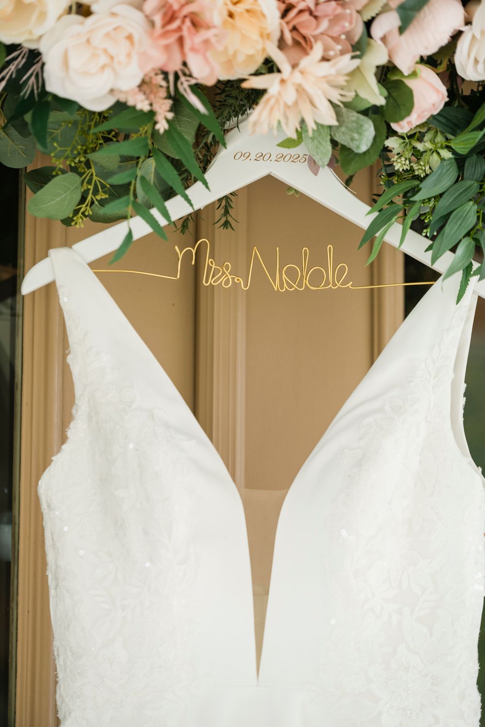 White lace wedding dress hanging on a custom Mrs. Noble hanger beneath pink and blush floral arrangement.