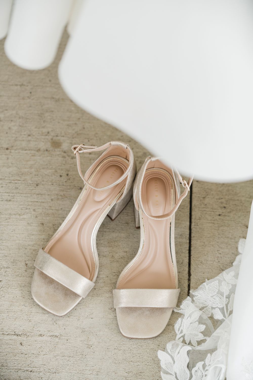 Elegant nude block heel sandals with ankle strap on wooden floor beside lace wedding dress.