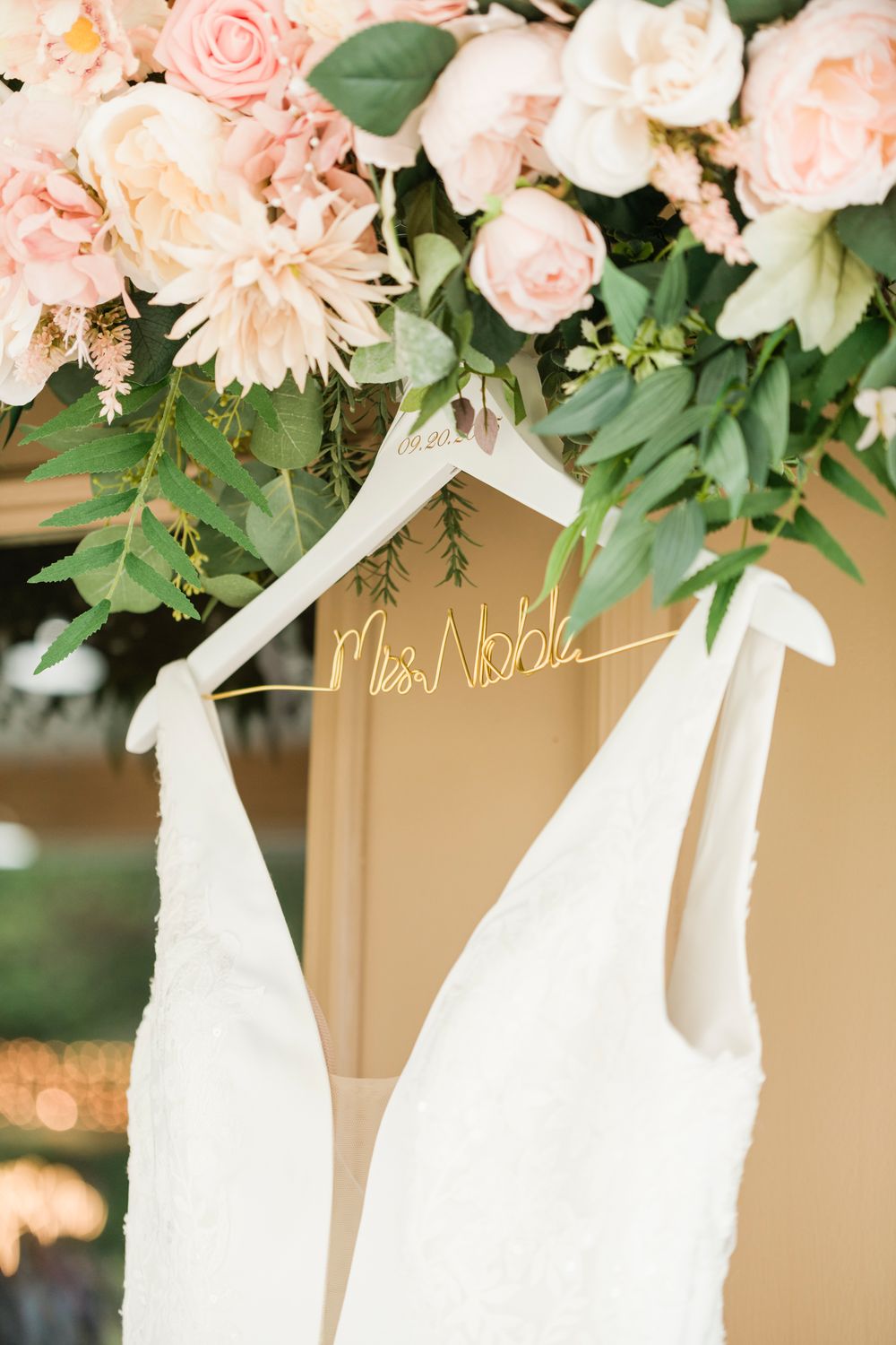 White wedding dress on personalized hanger reading Mrs. Noble, displayed beneath lush pink roses and greenery floral arrangement.