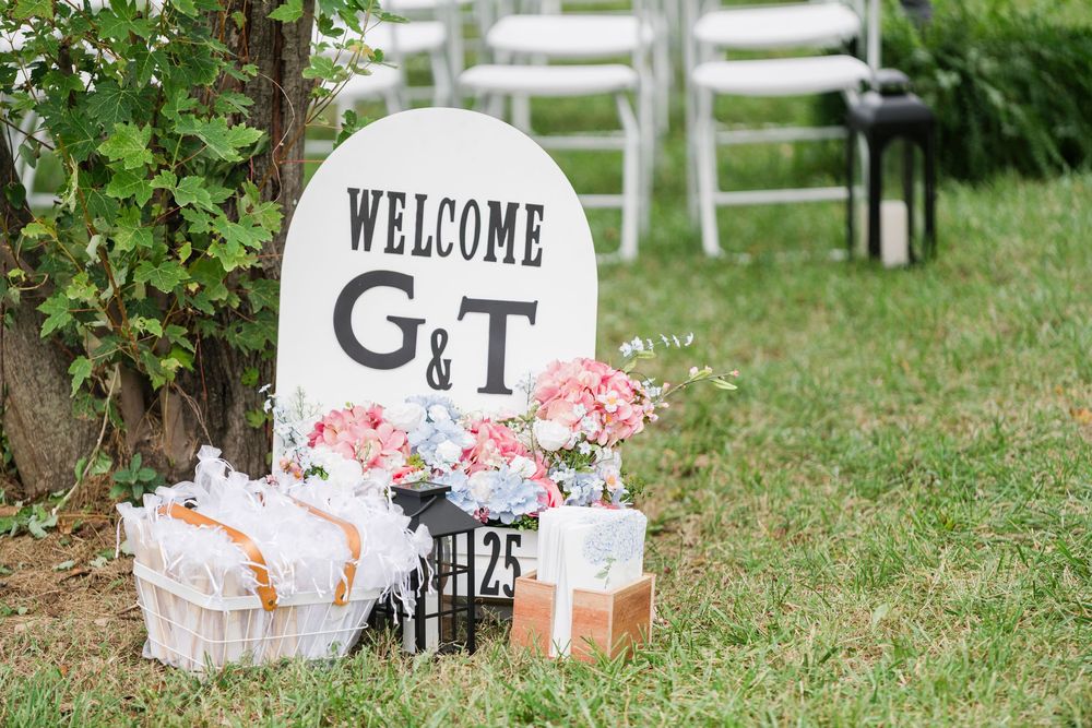 Outdoor wedding welcome sign with G&T monogram, pink floral arrangements, and gift baskets on green lawn.
