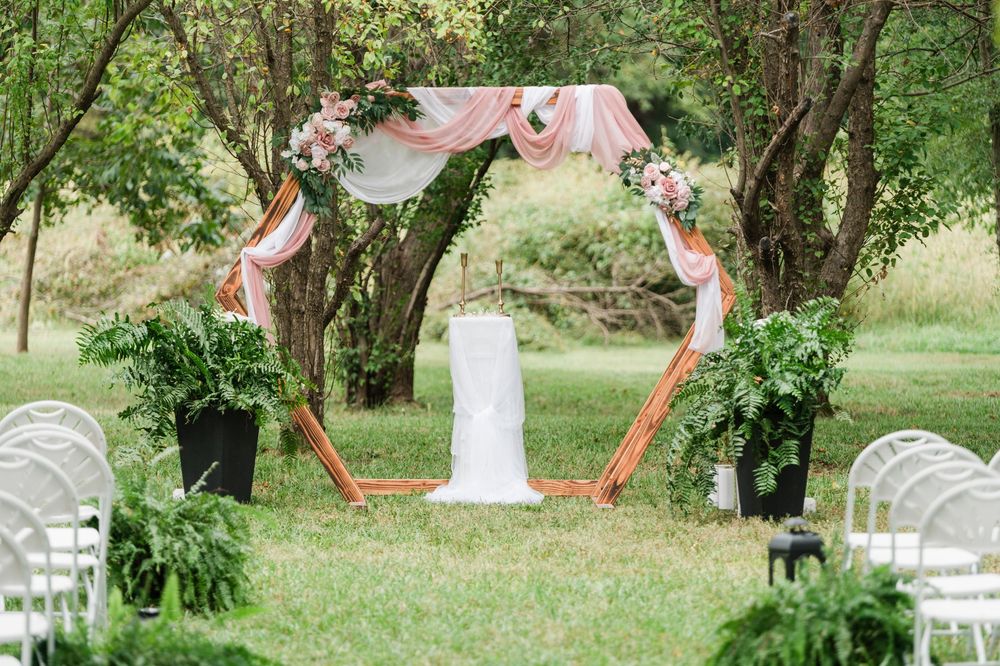 Outdoor wedding ceremony arch decorated with pink draping and flowers, surrounded by lush greenery and white chairs.