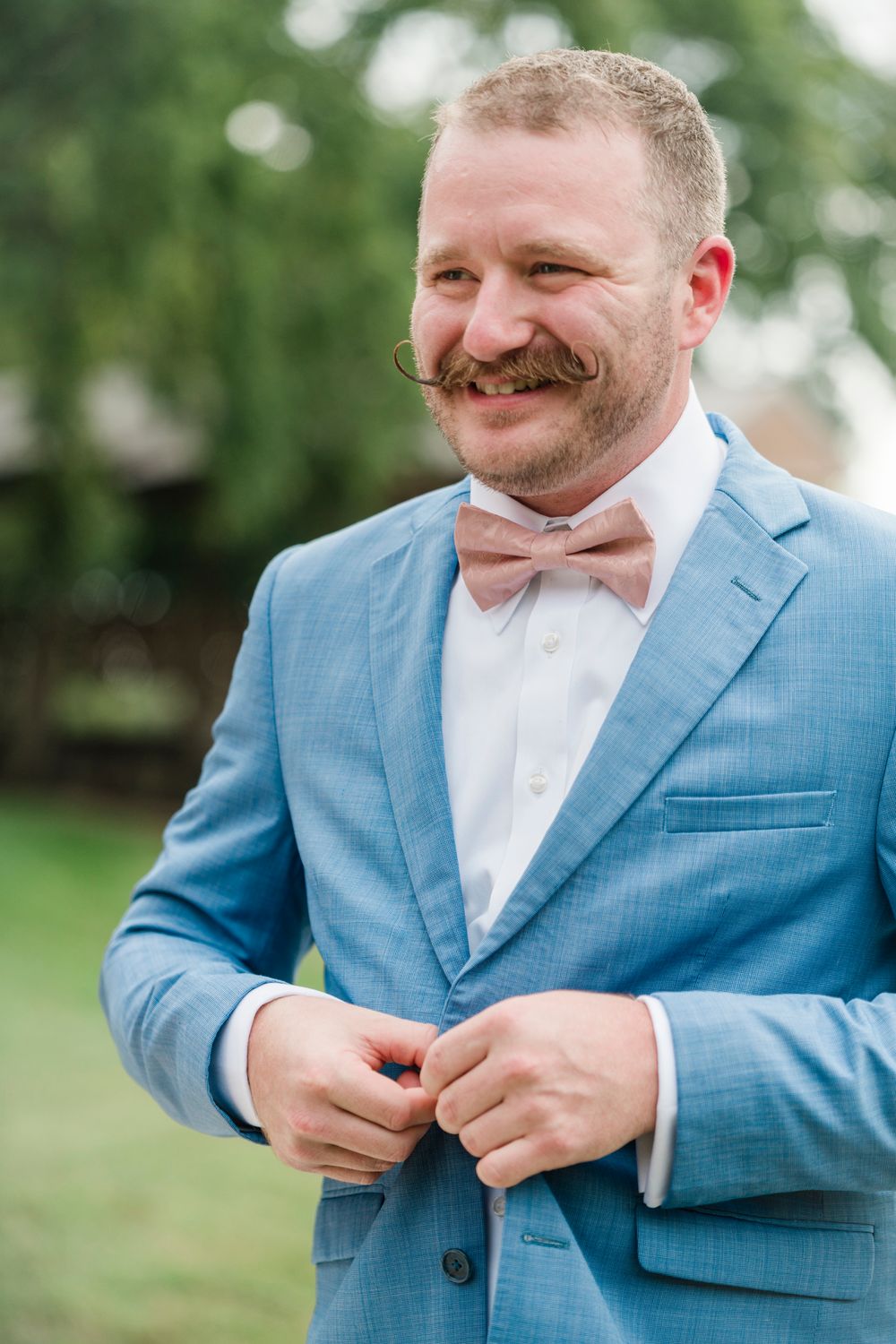 Smiling groom in blue suit and pink bow tie buttoning jacket outdoors.