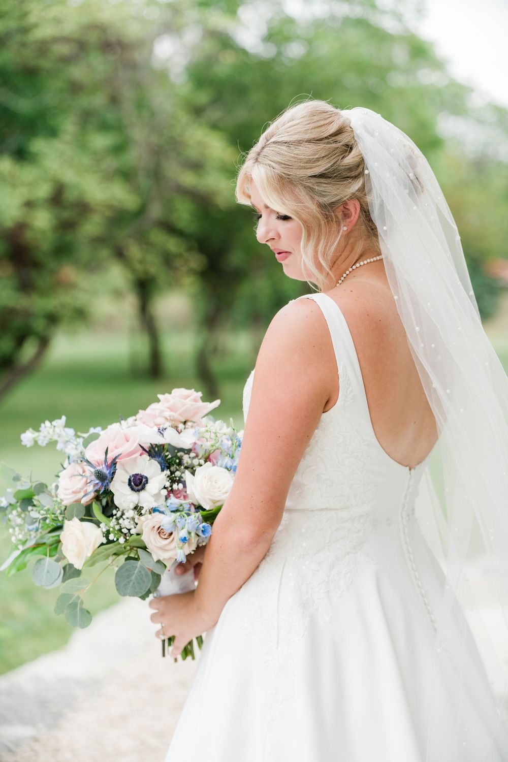 Bride in white gown and veil holds pink and blue bouquet outdoors, looking down gracefully.
