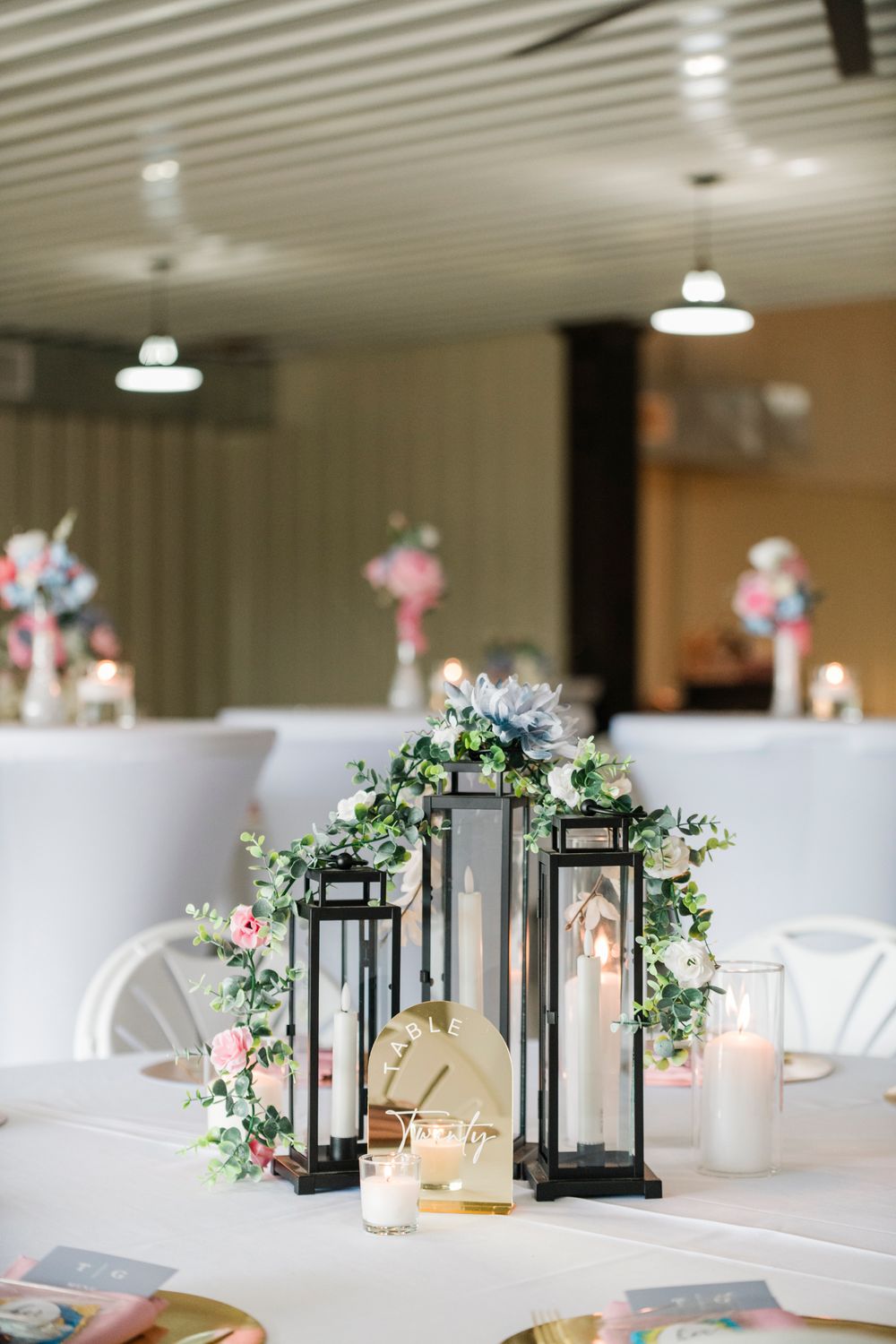 Elegant wedding reception table with black lantern centerpieces adorned with greenery and pink roses, white tablecloth, and candles.