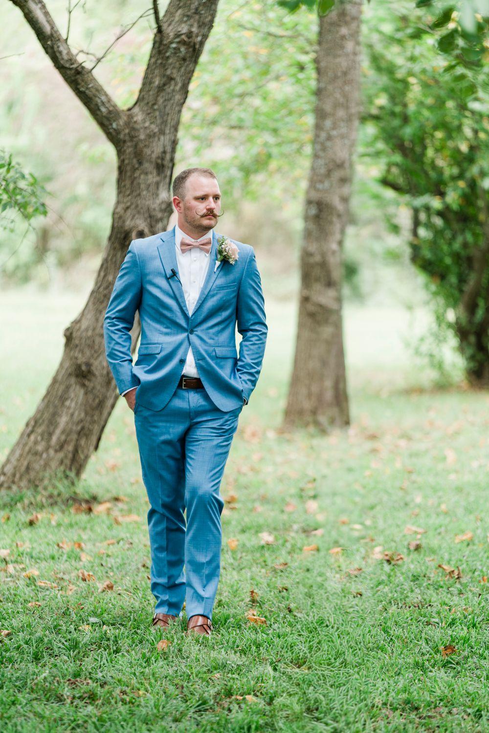 A groom in a blue suit with a floral boutonniere stands confidently on green grass among trees.