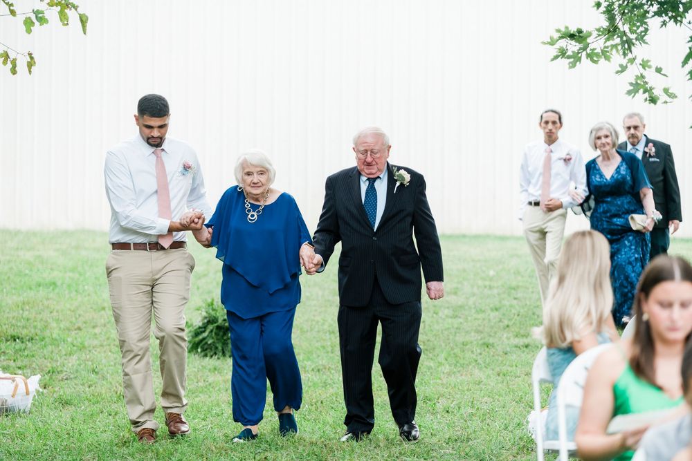 Elderly couple holding hands walks with young man at outdoor wedding ceremony on green lawn.