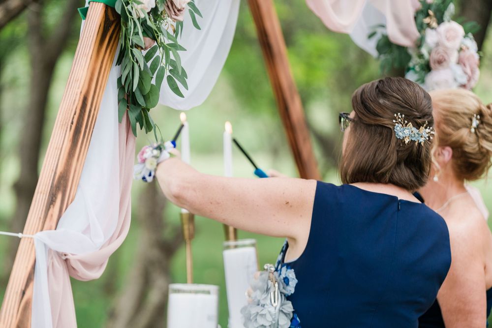 Two women lighting unity candles at an outdoor wedding ceremony decorated with floral arch and flowing fabric.