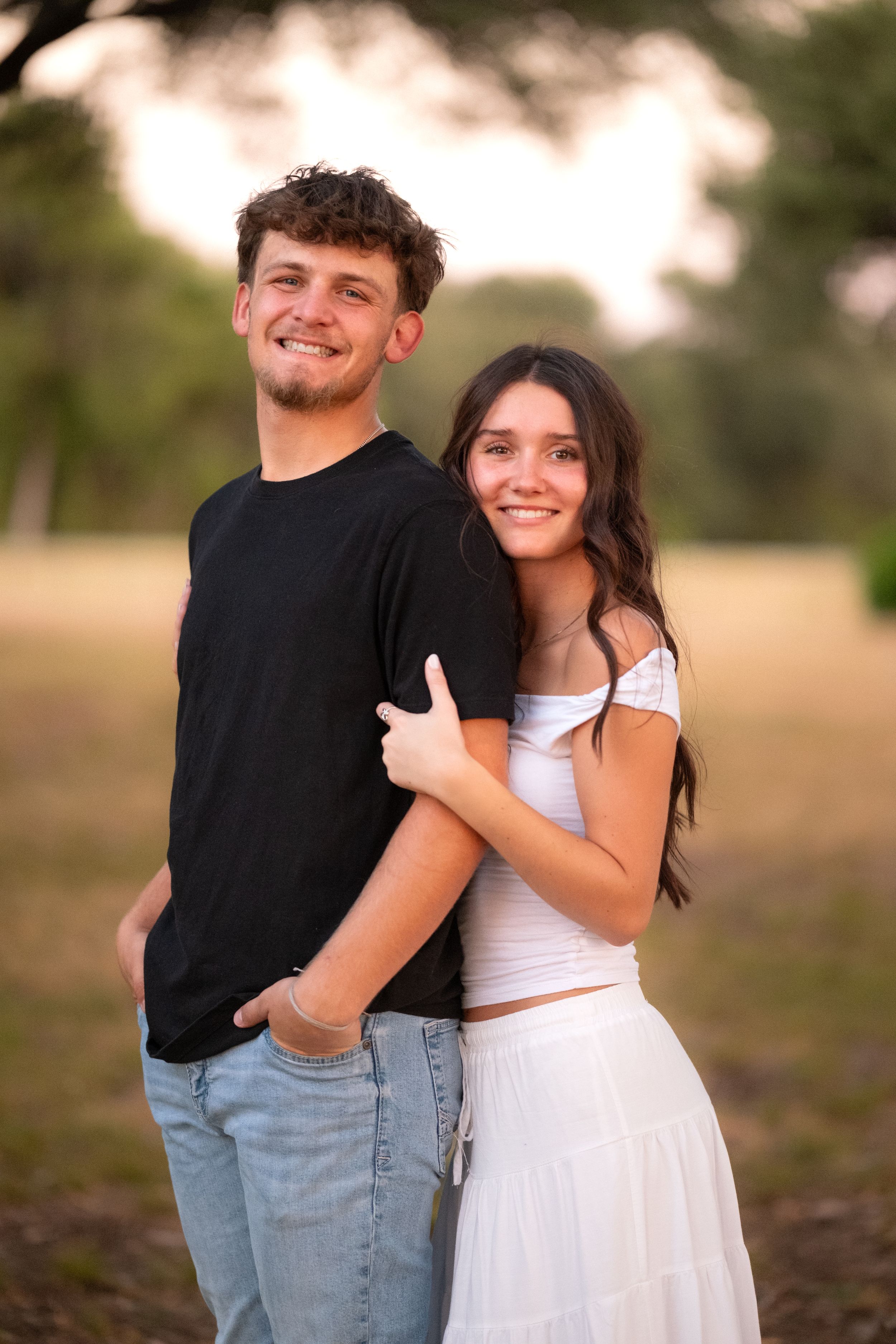Young couple smiling and embracing in a sunlit park, woman in white dress hugging man in black tee in Belton Texas
