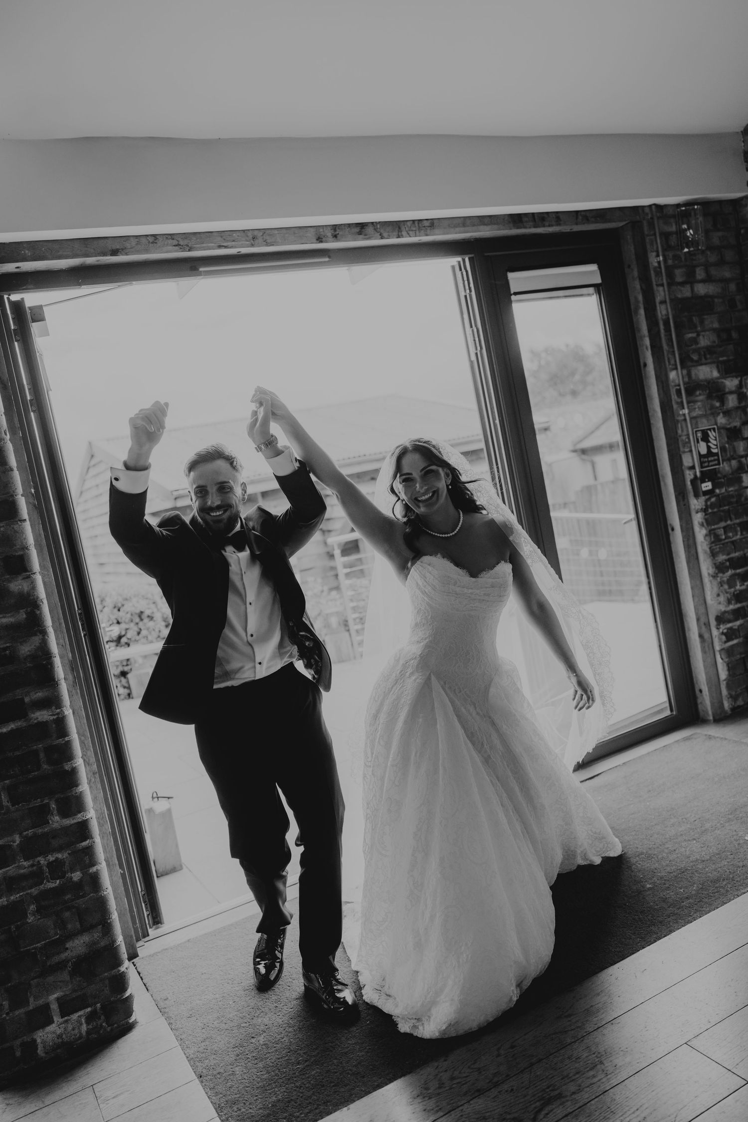 Black and white photo of joyful newlyweds celebrating their grand entrance, hands raised, bride in gown, groom in suit.