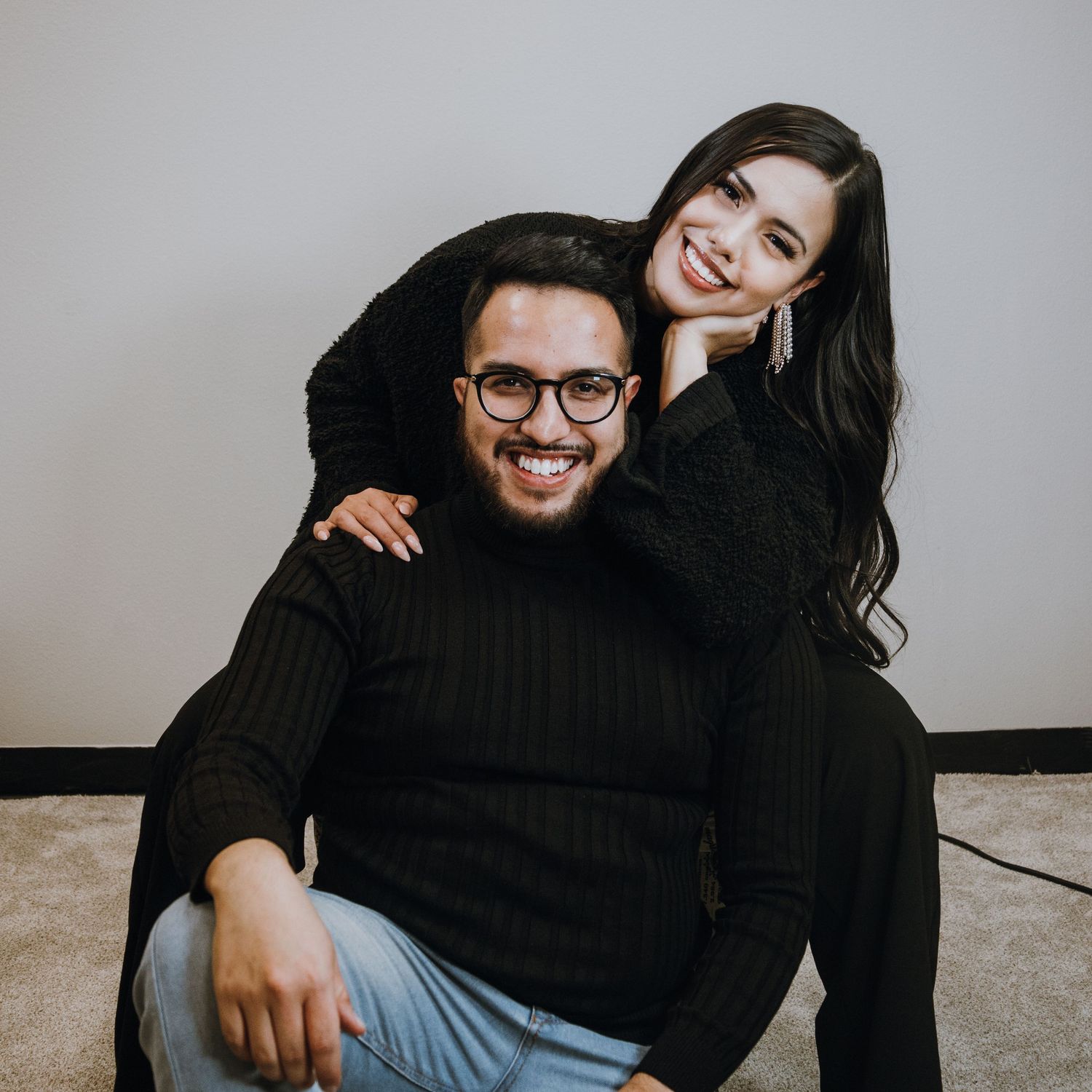 Smiling couple in black outfits posing together playfully against a neutral gray background.