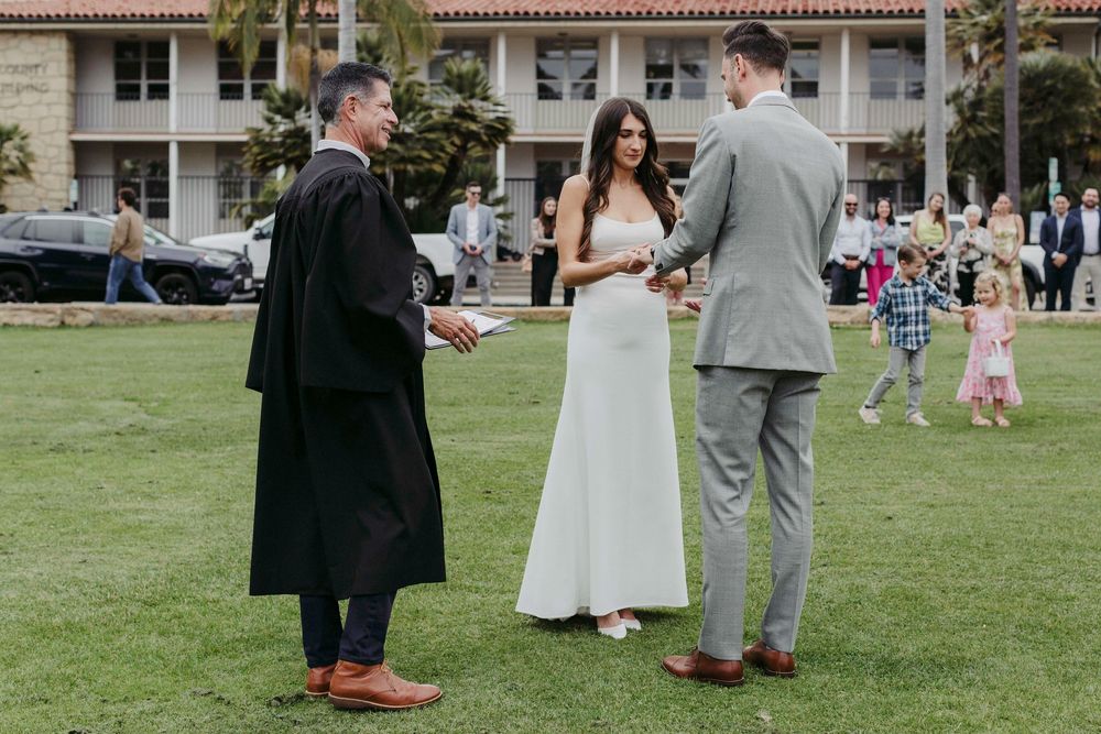 Outdoor wedding ceremony on green lawn with officiant in black robe, bride in white dress, and groom in gray suit.