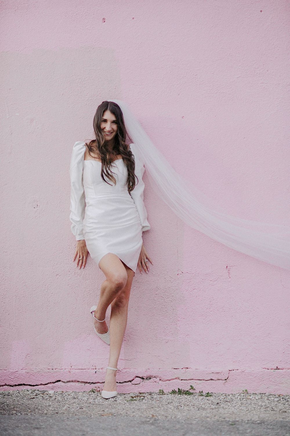 Bride in short white dress and veil poses against pink wall, one leg raised, smiling.