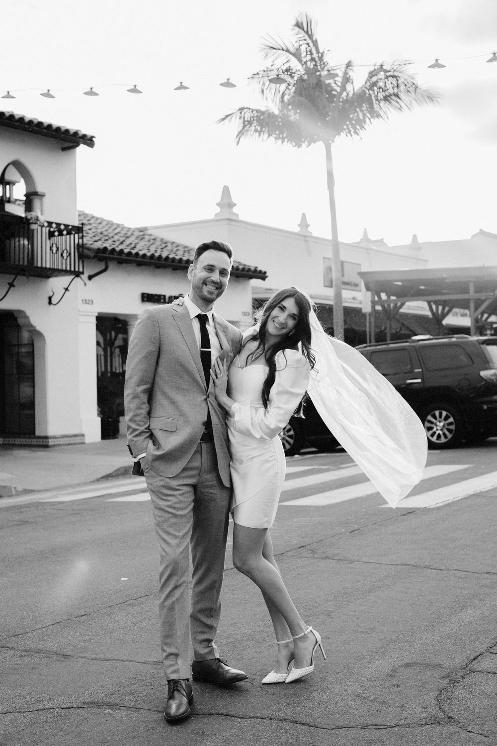 Black and white photo of a smiling couple in wedding attire posing outdoors near a palm tree and Spanish-style building.