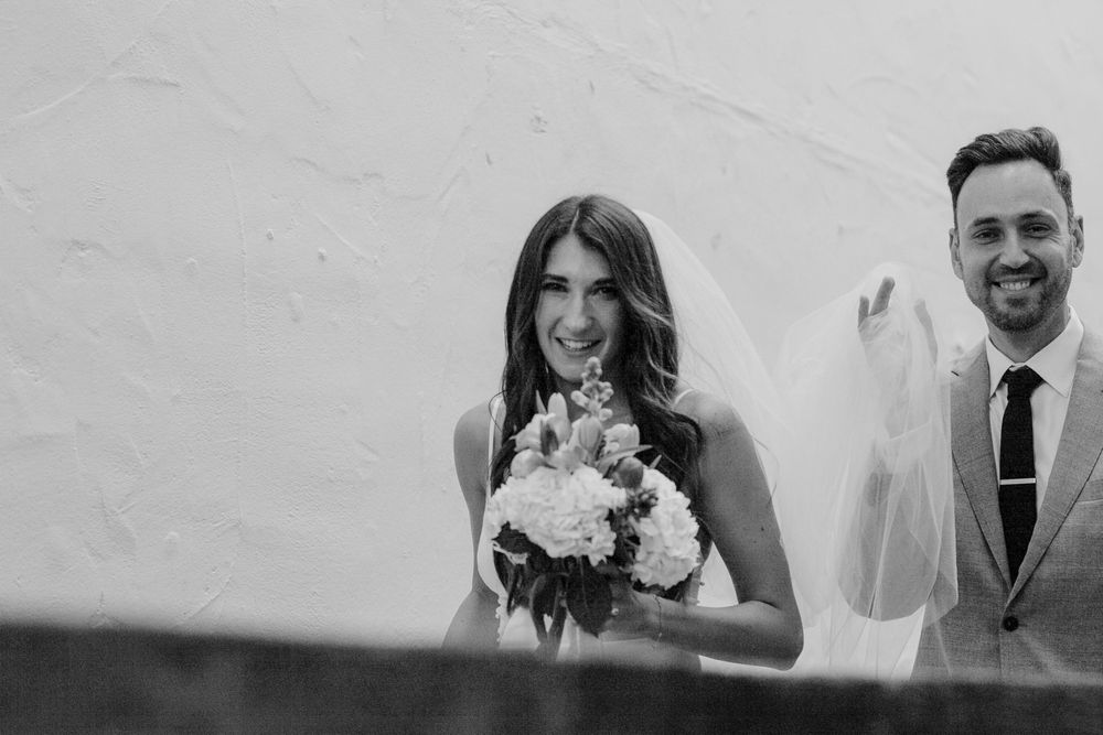 Black and white photo of a smiling bride holding a bouquet with a groom in a suit waving behind her.
