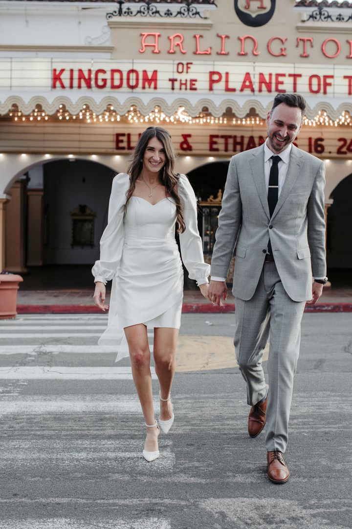 A bride and groom hold hands crossing a street in front of the Arlington Theatre marquee on their wedding day.