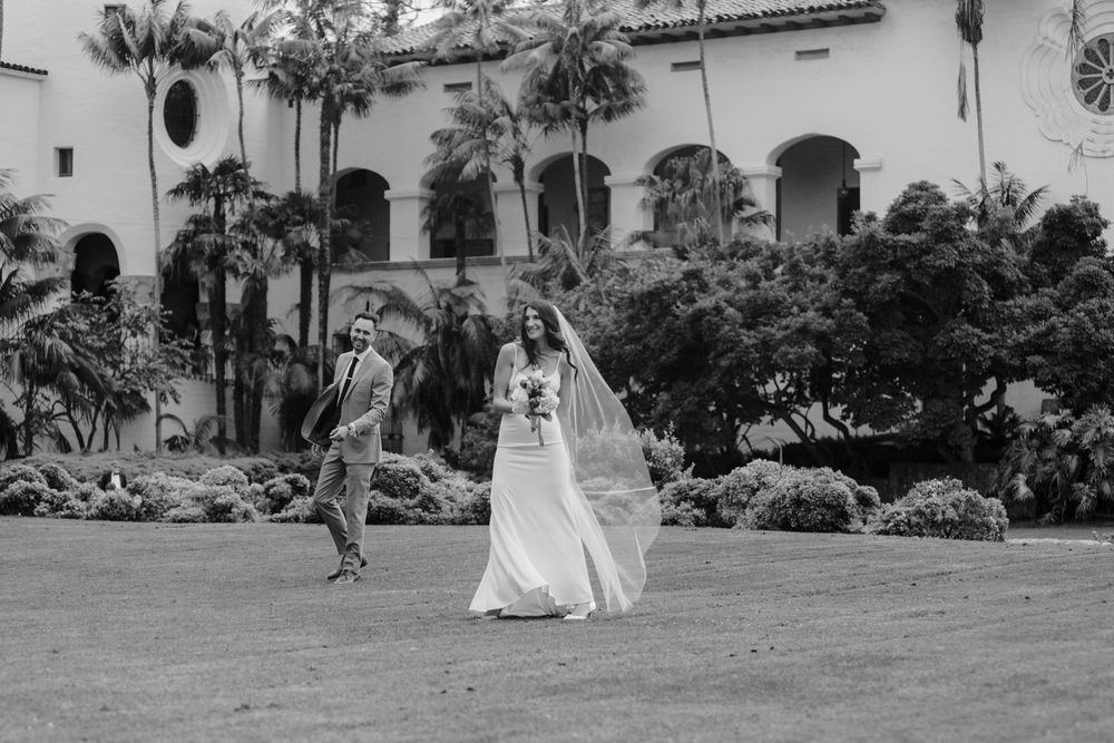 Black and white photo of a bride and groom walking on a lawn in front of an elegant building with palm trees.