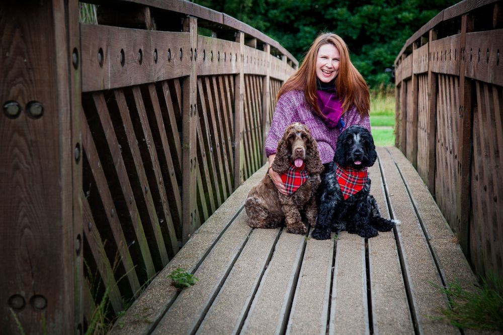 Woman bent down hugging her two spaniels that are wearing red bandanas. The woman is also wearing a handmade neck scarf.