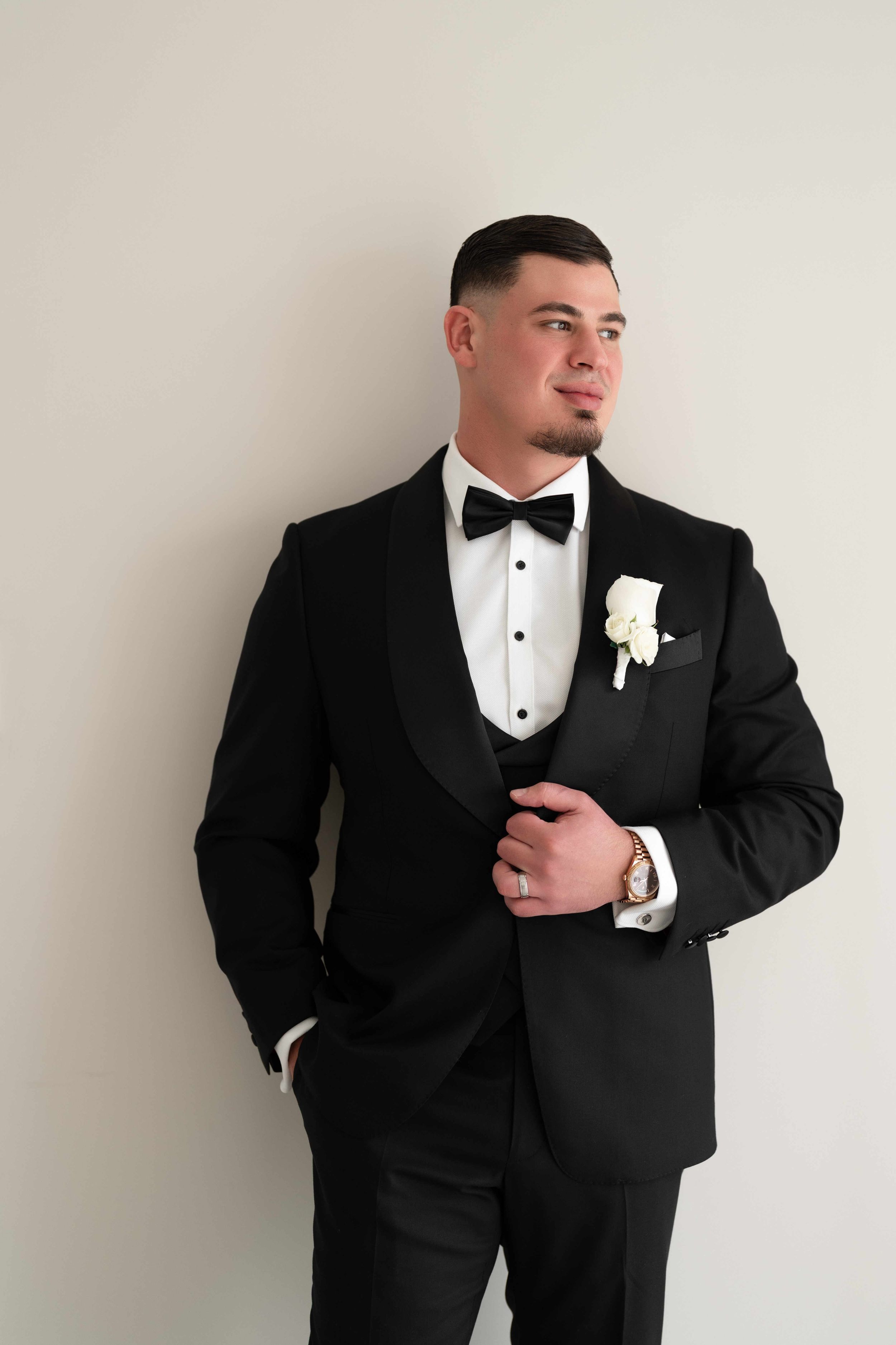 A groom in a black tuxedo with bow tie and white boutonniere poses confidently against a light gray wall.