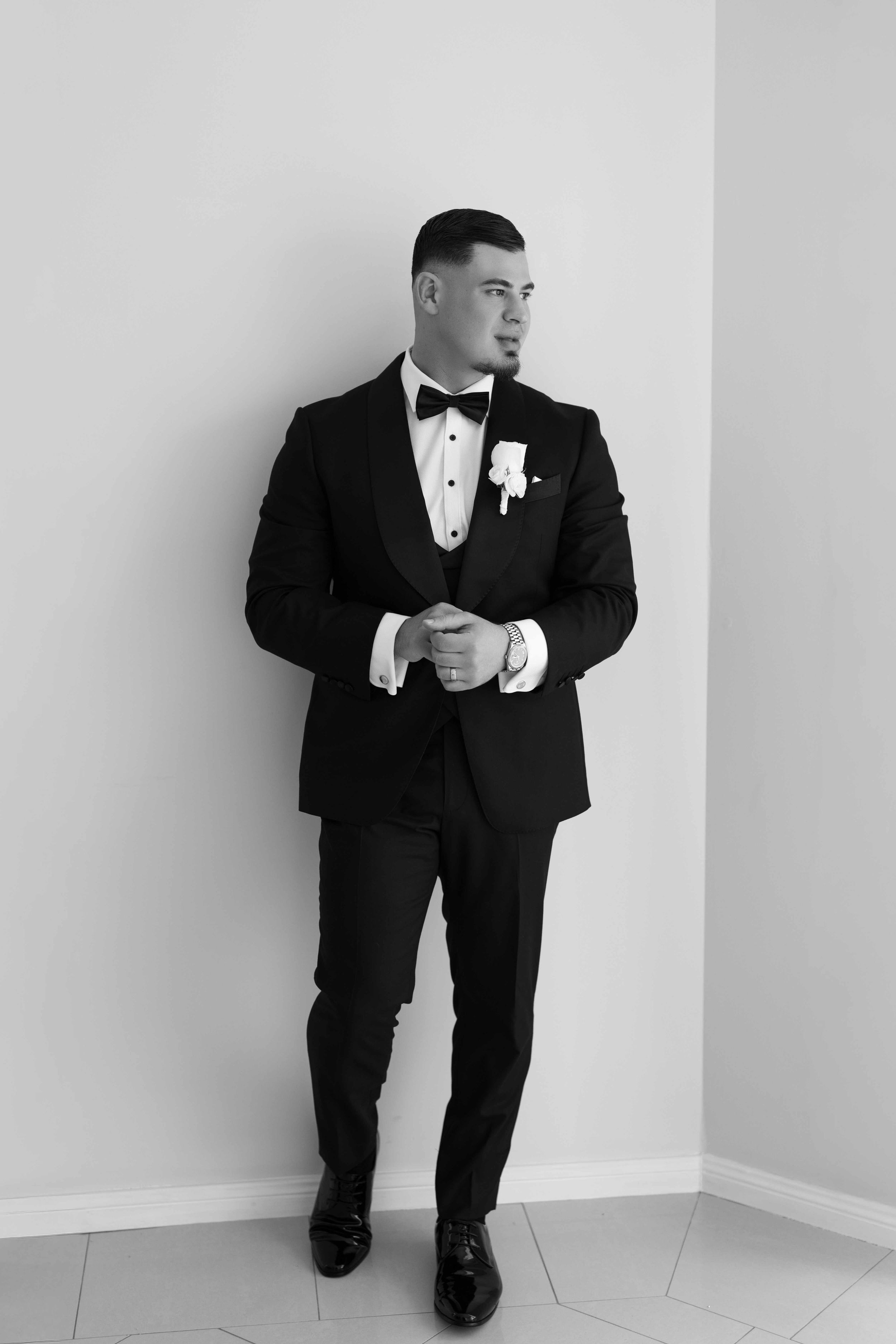 Groom in black tuxedo with bow tie and boutonniere poses against white wall in elegant black and white portrait.