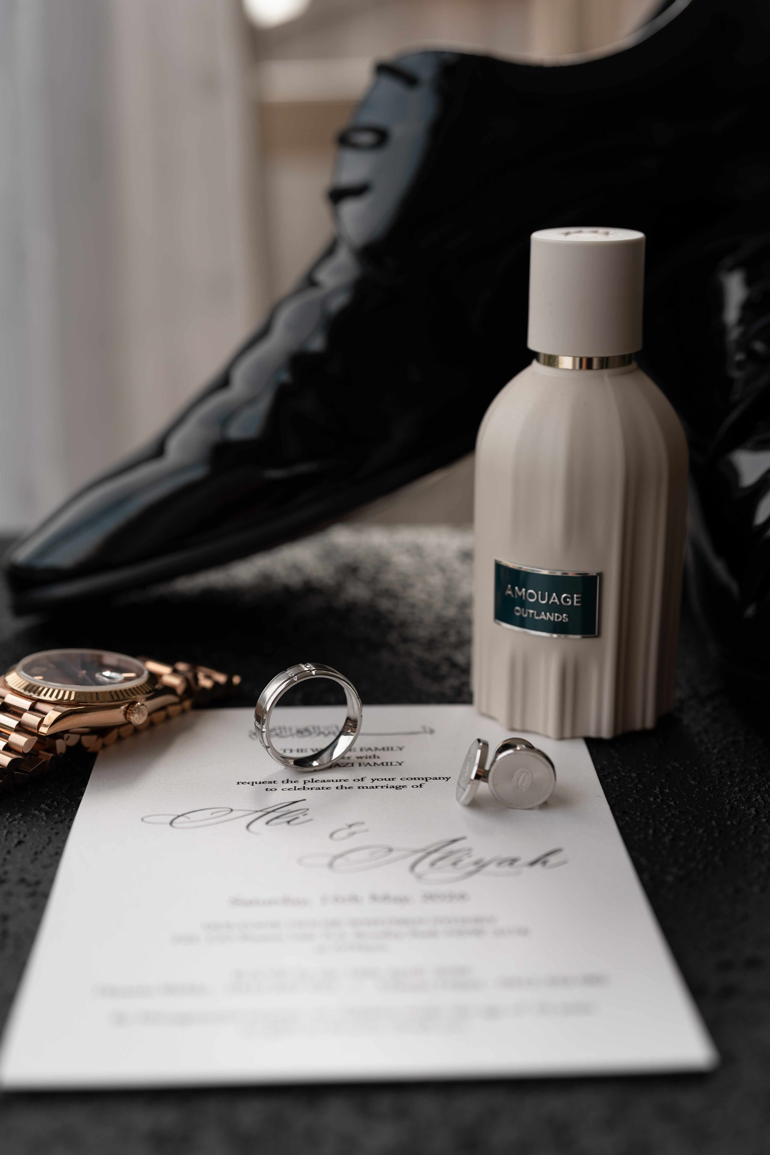 Elegant groom wedding flat lay featuring Amouage cologne, gold watch, wedding rings, cufflinks, invitation, and black dress shoes.
