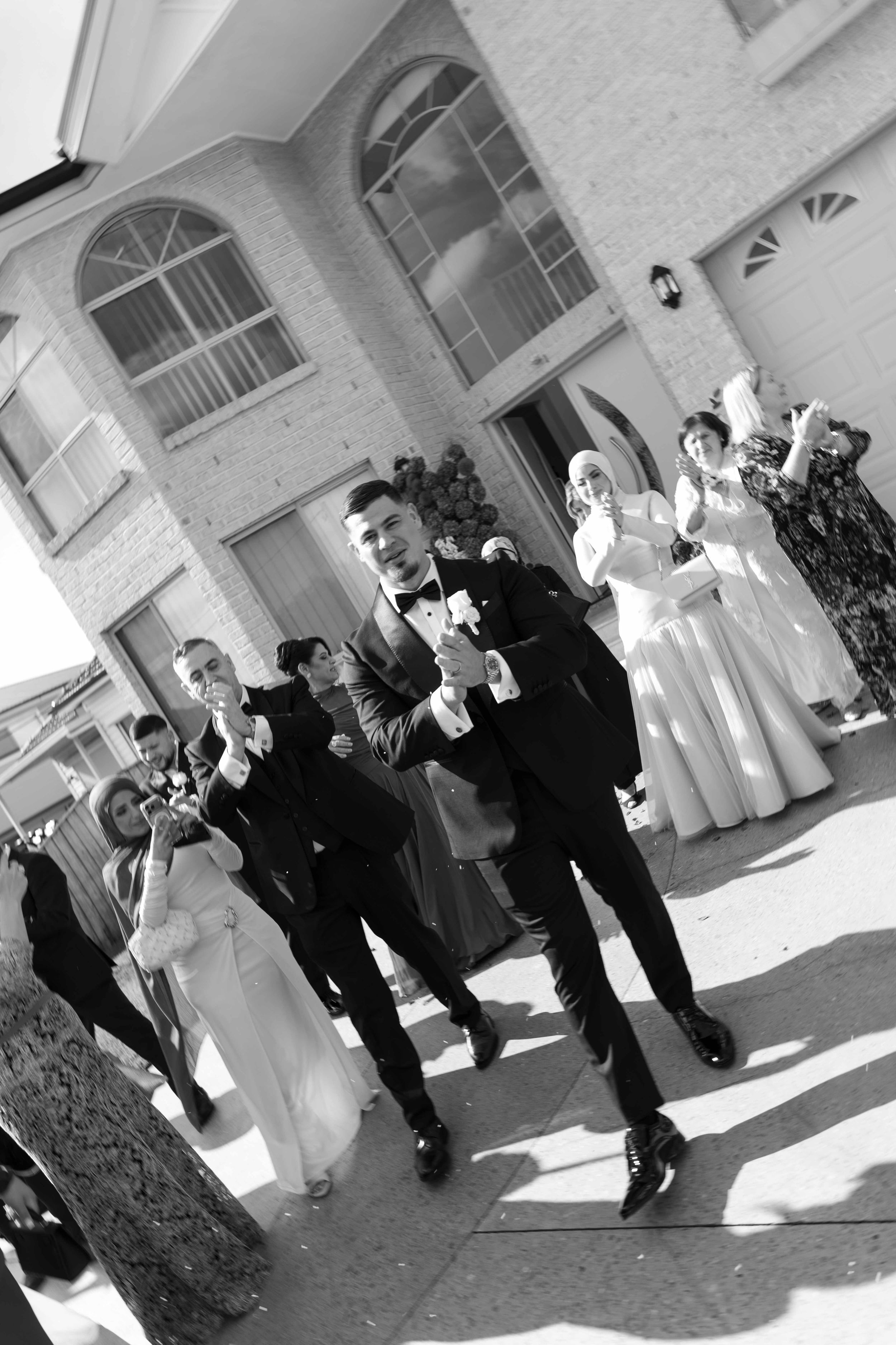 Black and white photo of groom and groomsmen celebrating outside a brick house during a wedding ceremony.