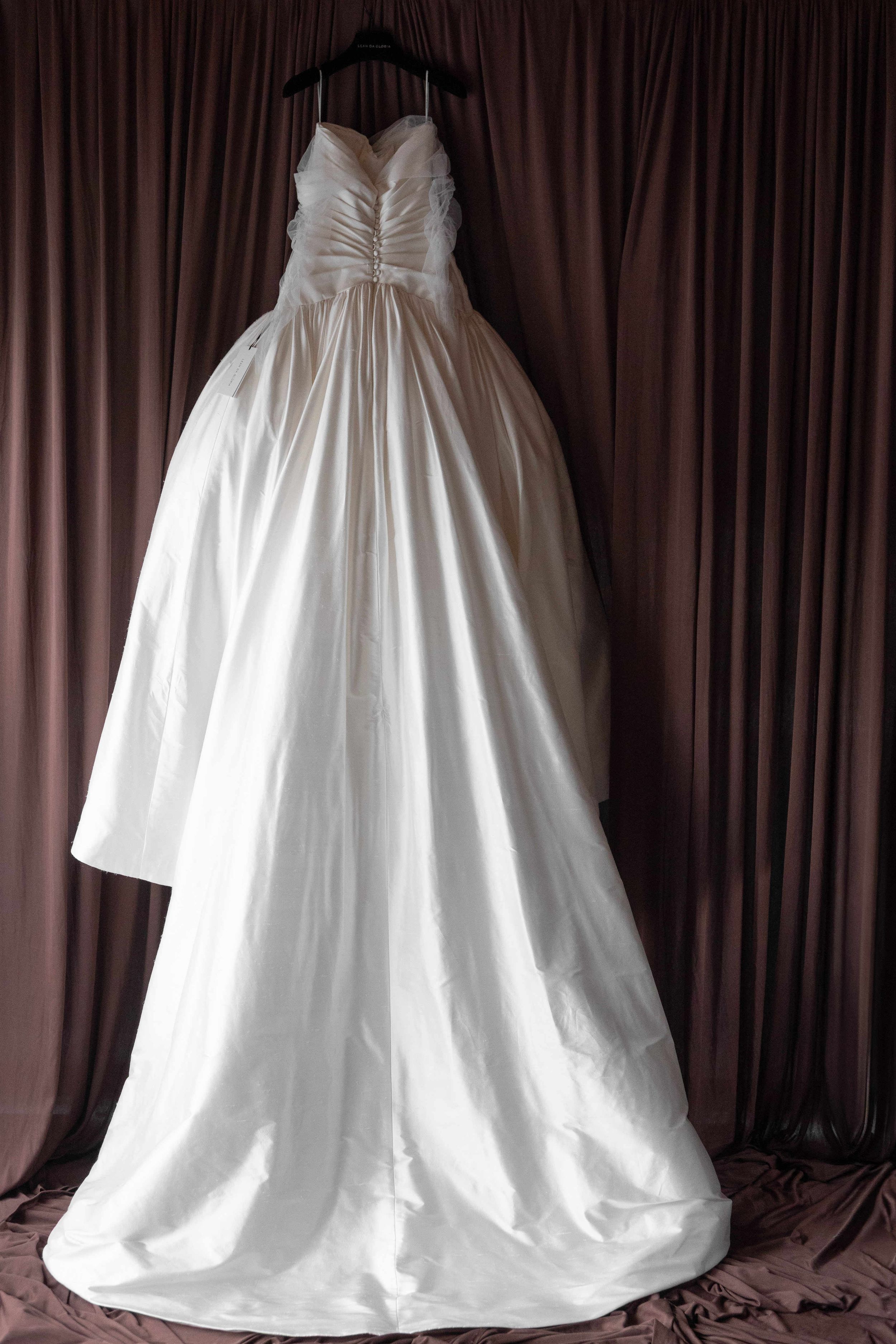 Elegant white strapless ball gown wedding dress with ruched bodice hanging on a hanger against brown drape backdrop.