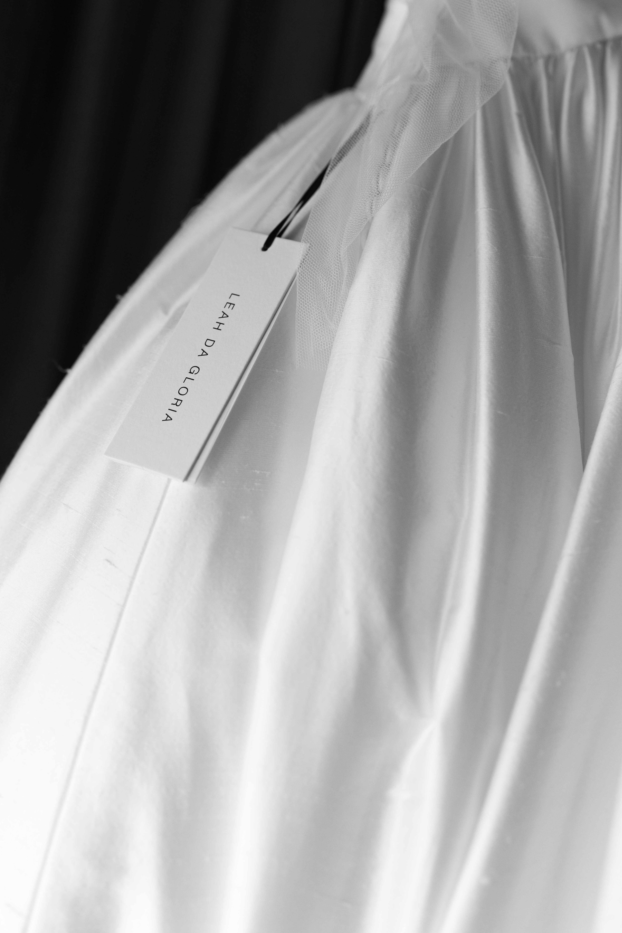 Black and white photo of a white bridal gown on a hanger with a Leah Da Gloria designer tag.