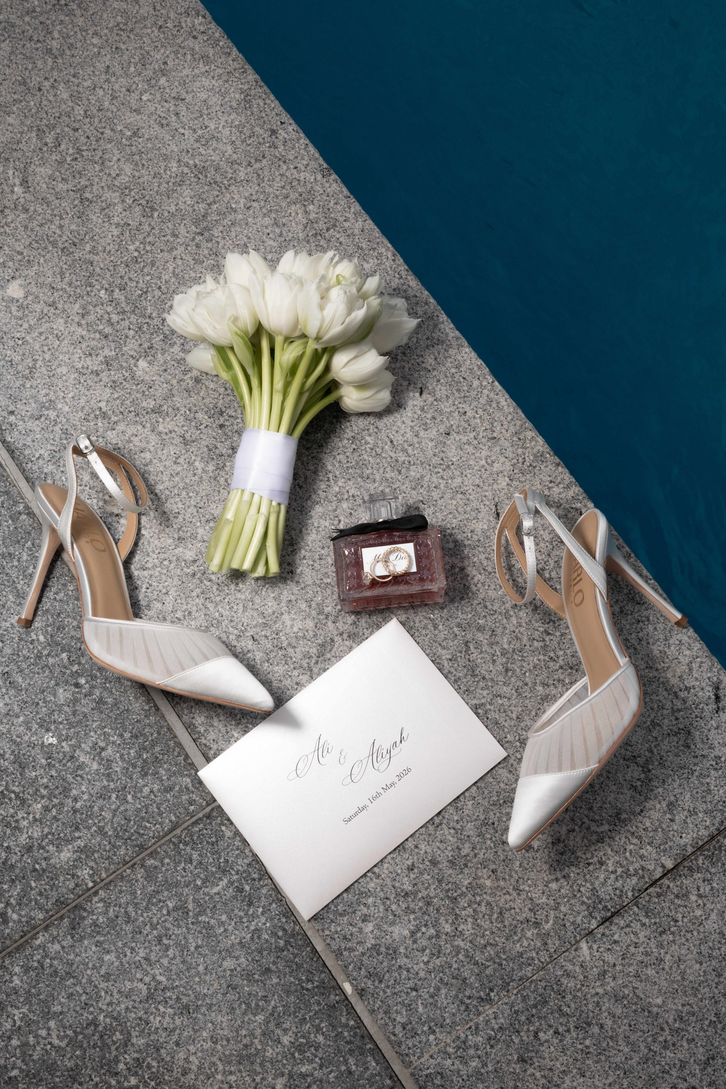 Bridal flat lay with white tulip bouquet, high heel sandals, wedding invitation, and perfume bottle by a pool.