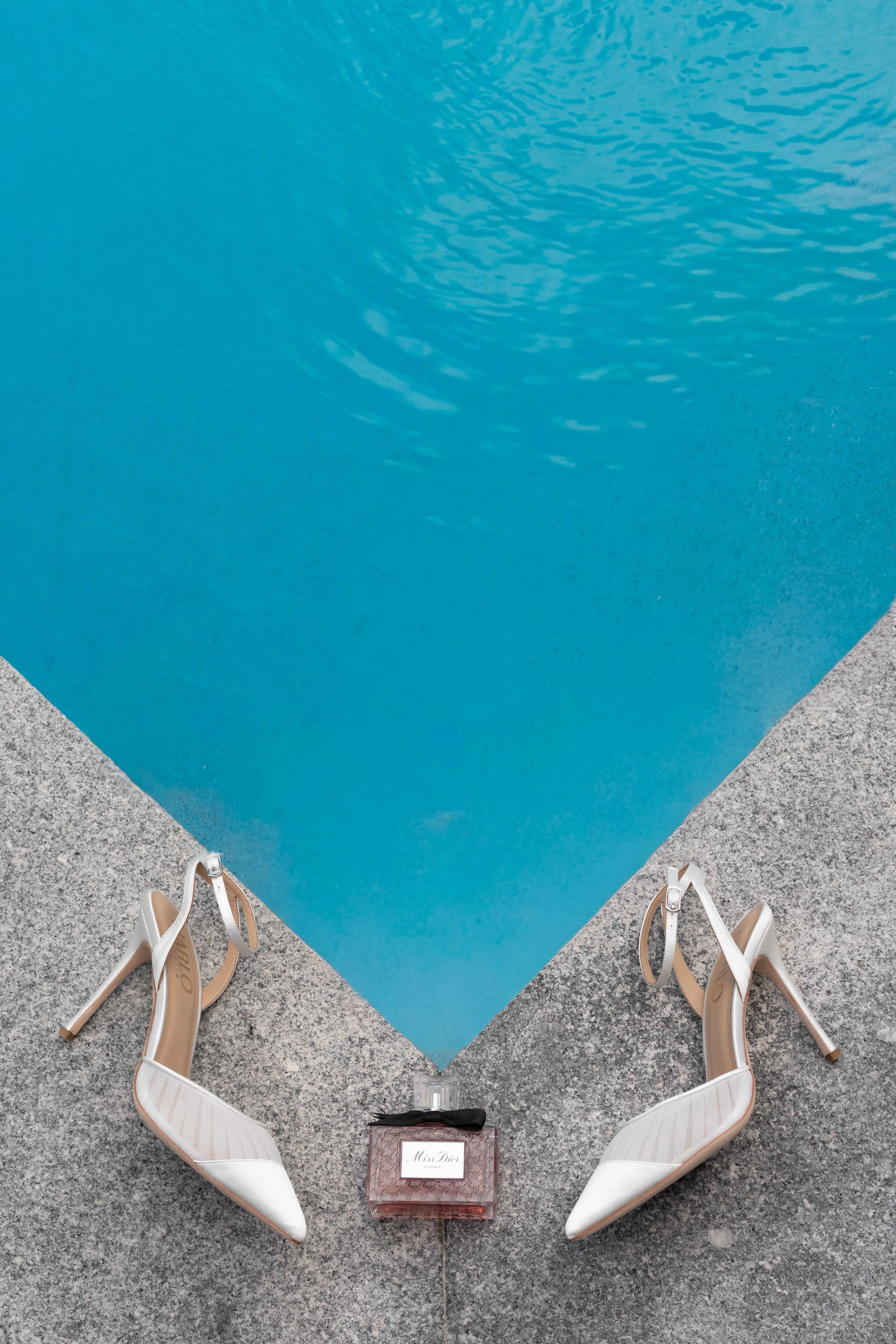 White slingback heels and a perfume bottle arranged on pool edge with turquoise water, elegant bridal accessories flatlay.