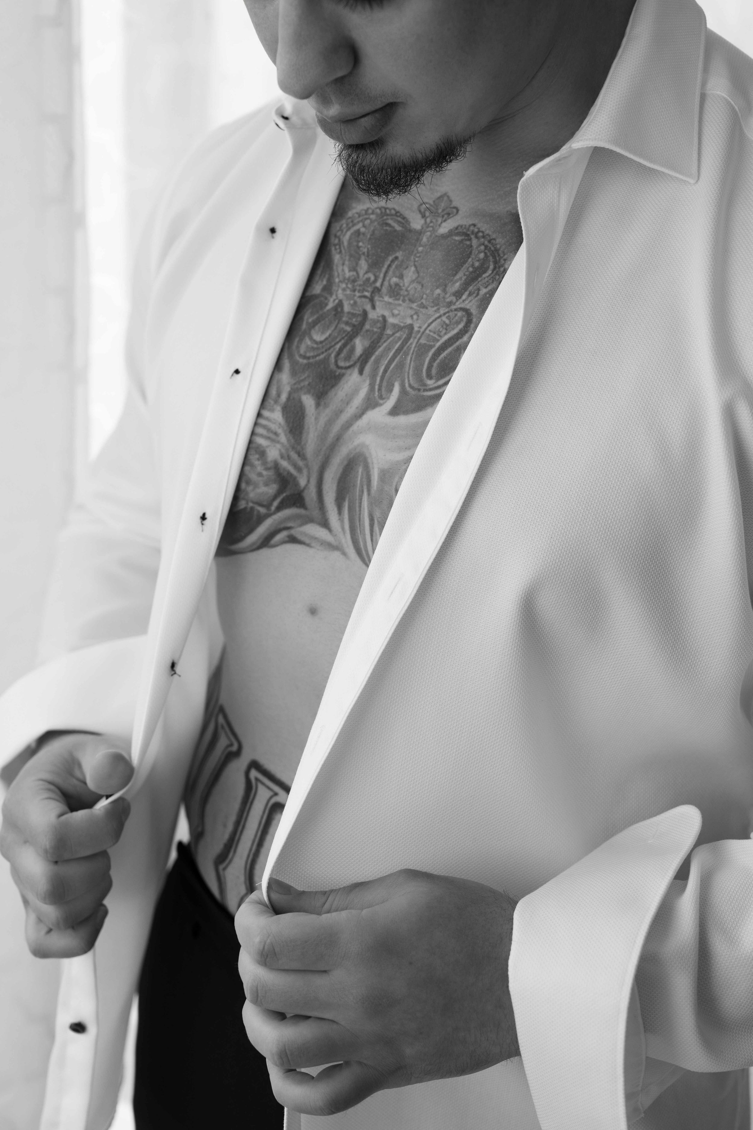 Black and white photo of a tattooed man buttoning a white dress shirt, revealing chest tattoos.