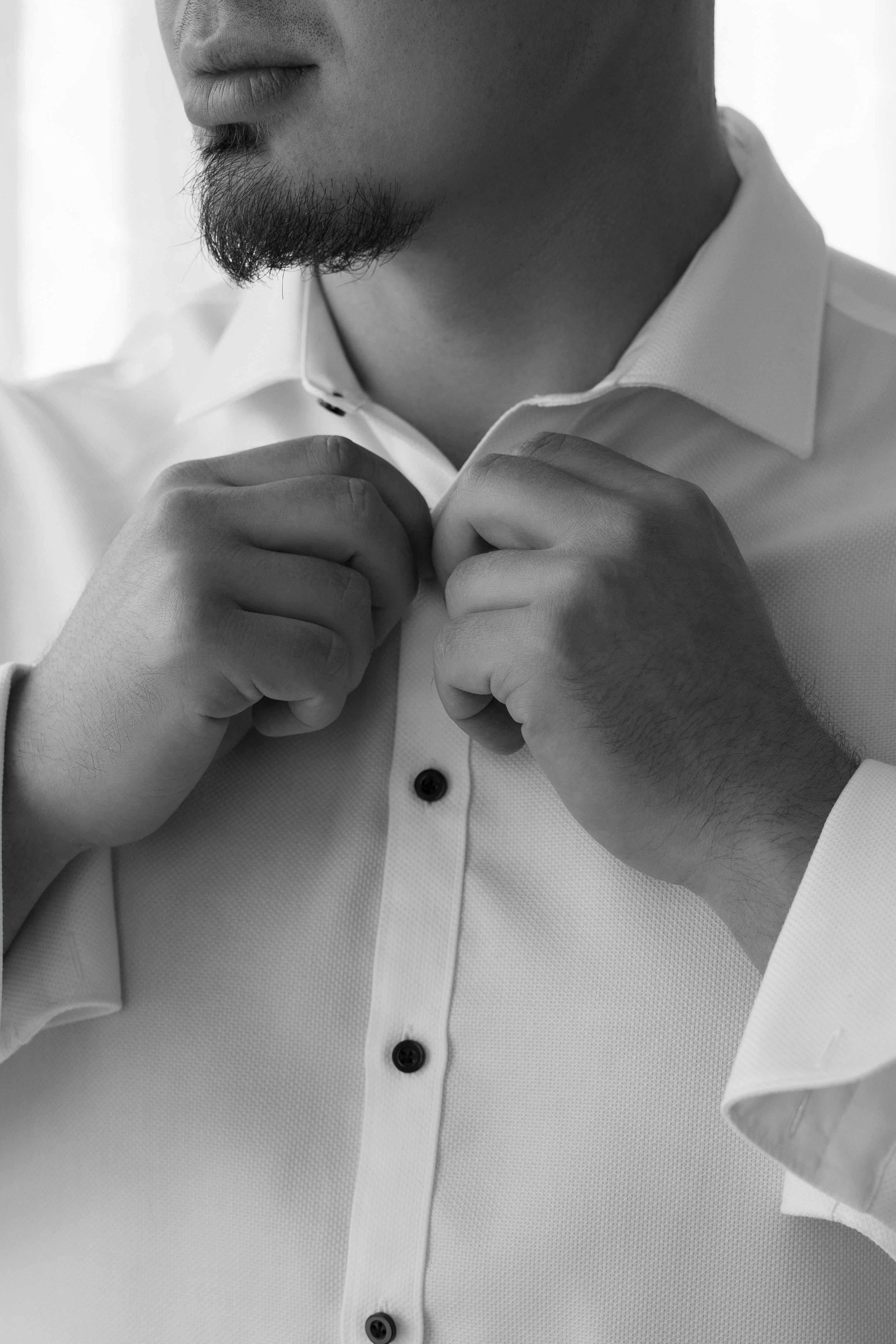 Black and white photo of a man buttoning up a white dress shirt, preparing for a formal occasion.