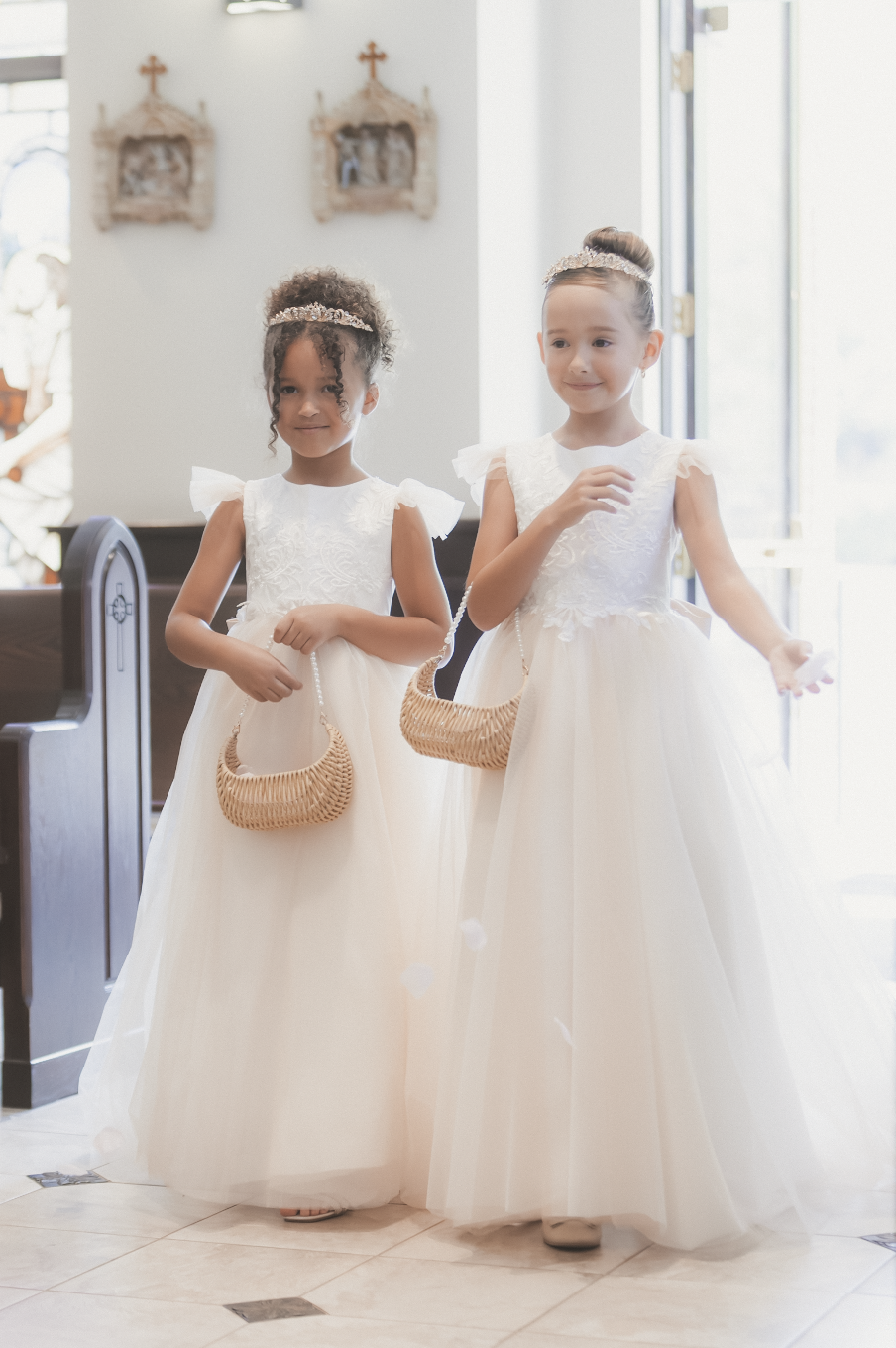 Two flower girls in white tulle dresses with wicker baskets pose in a church aisle.