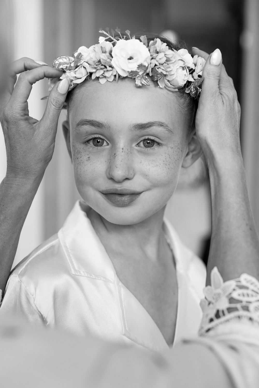 Black and white photo of a young girl with freckles having a floral crown placed on her head.