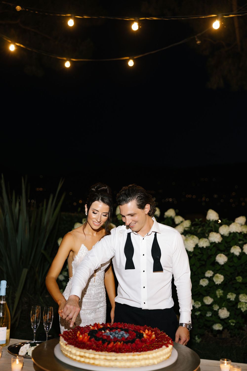 Bride and groom cutting a berry tart wedding cake outdoors at night under string lights.