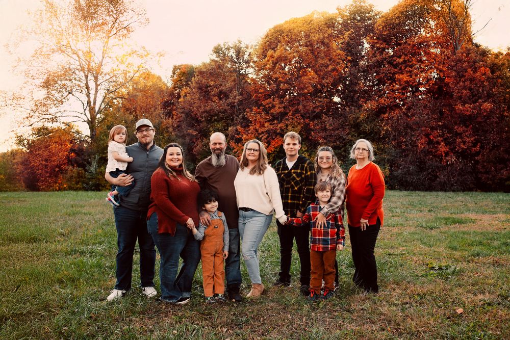 Family portrait of nine people posing in a field with vibrant autumn foliage in warm golden hour light.