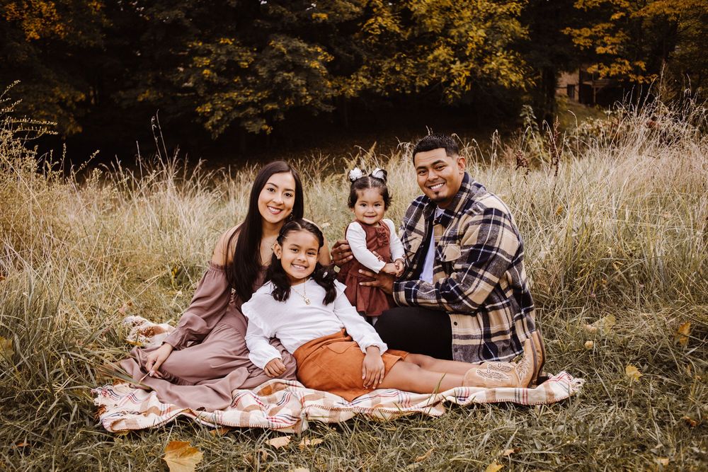 Family of four smiling together on a blanket in an autumn field with golden grass and fall foliage.