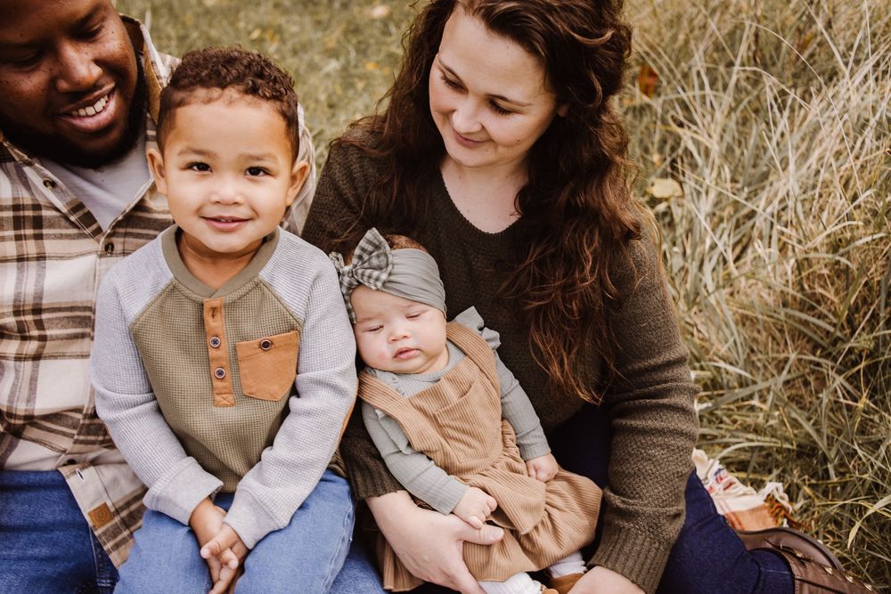 A happy interracial family of four sits outdoors in autumn grass, parents holding a toddler and newborn baby girl.