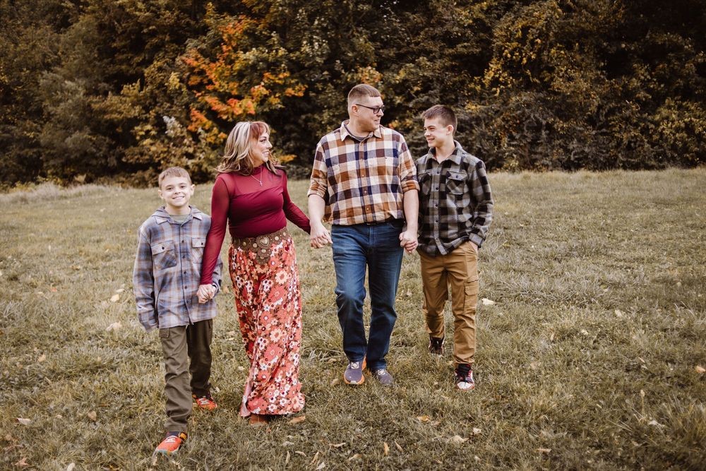 Family of four walks through autumn field, parents holding hands with two boys in flannel shirts amid fall foliage.
