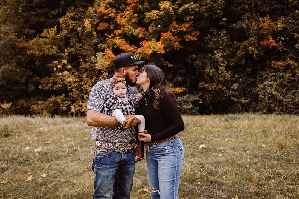 A couple kisses while holding their baby outdoors amid vibrant fall foliage in a scenic meadow.