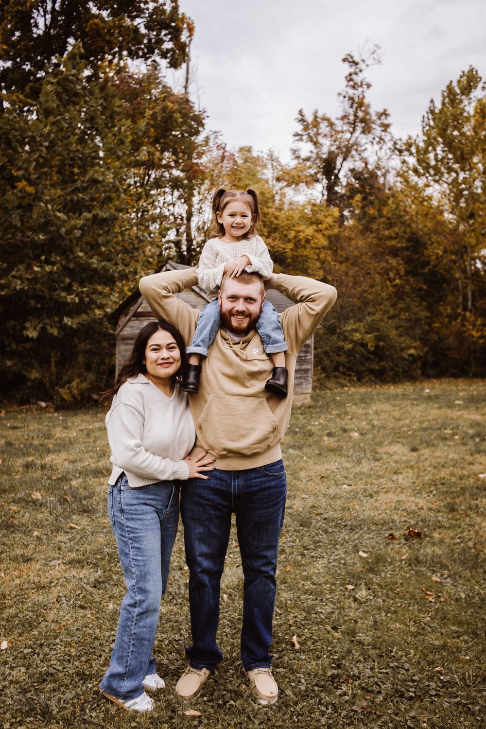A happy family of three poses outdoors in autumn, with a young girl sitting on her father's shoulders beside her smiling mother.