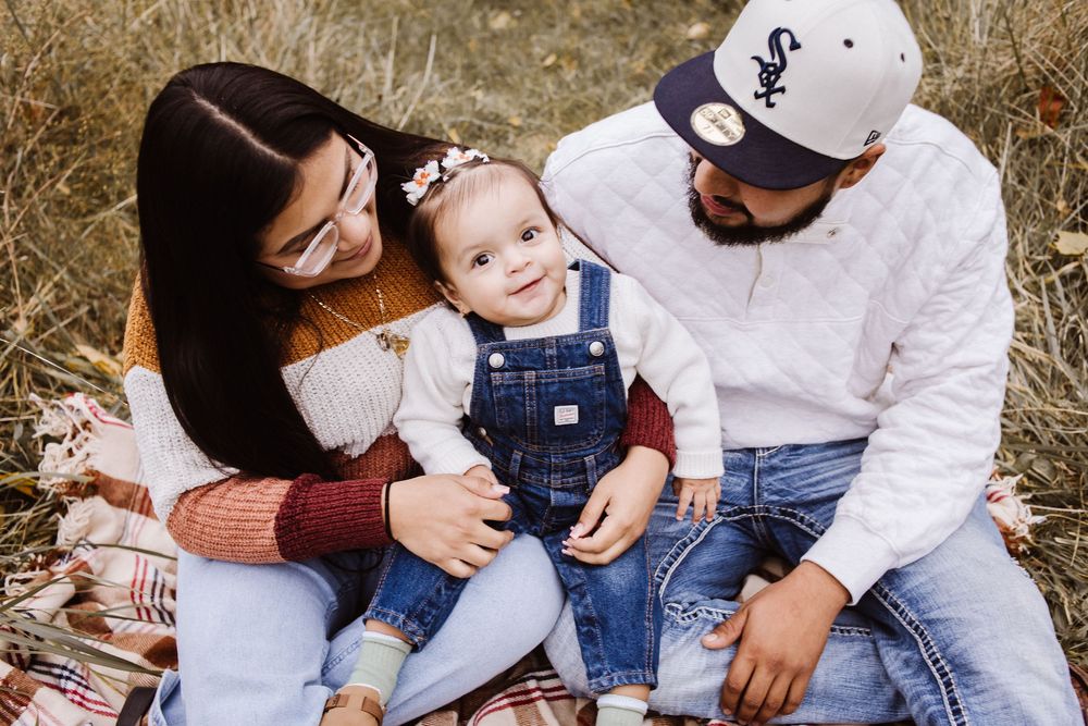 A young family sits outdoors on a blanket, smiling at their baby in denim overalls during a fall photo session.