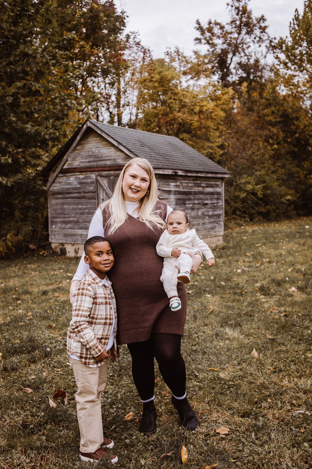 A woman in a brown dress holds a baby while standing with a young boy outdoors near a rustic shed in autumn.
