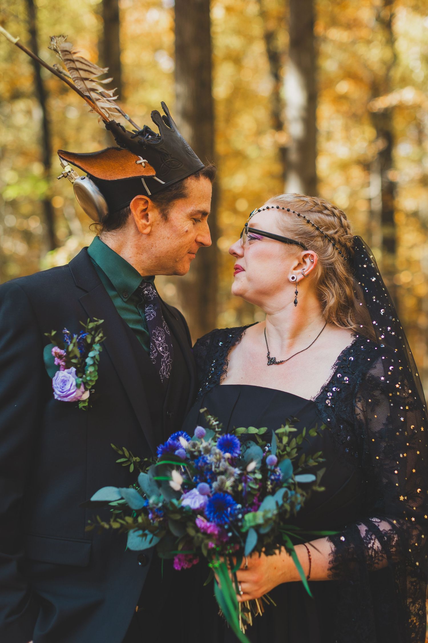 Gothic wedding couple in autumn forest, bride in black dress with veil, groom wearing decorative horned hat, holding blue bouquet.