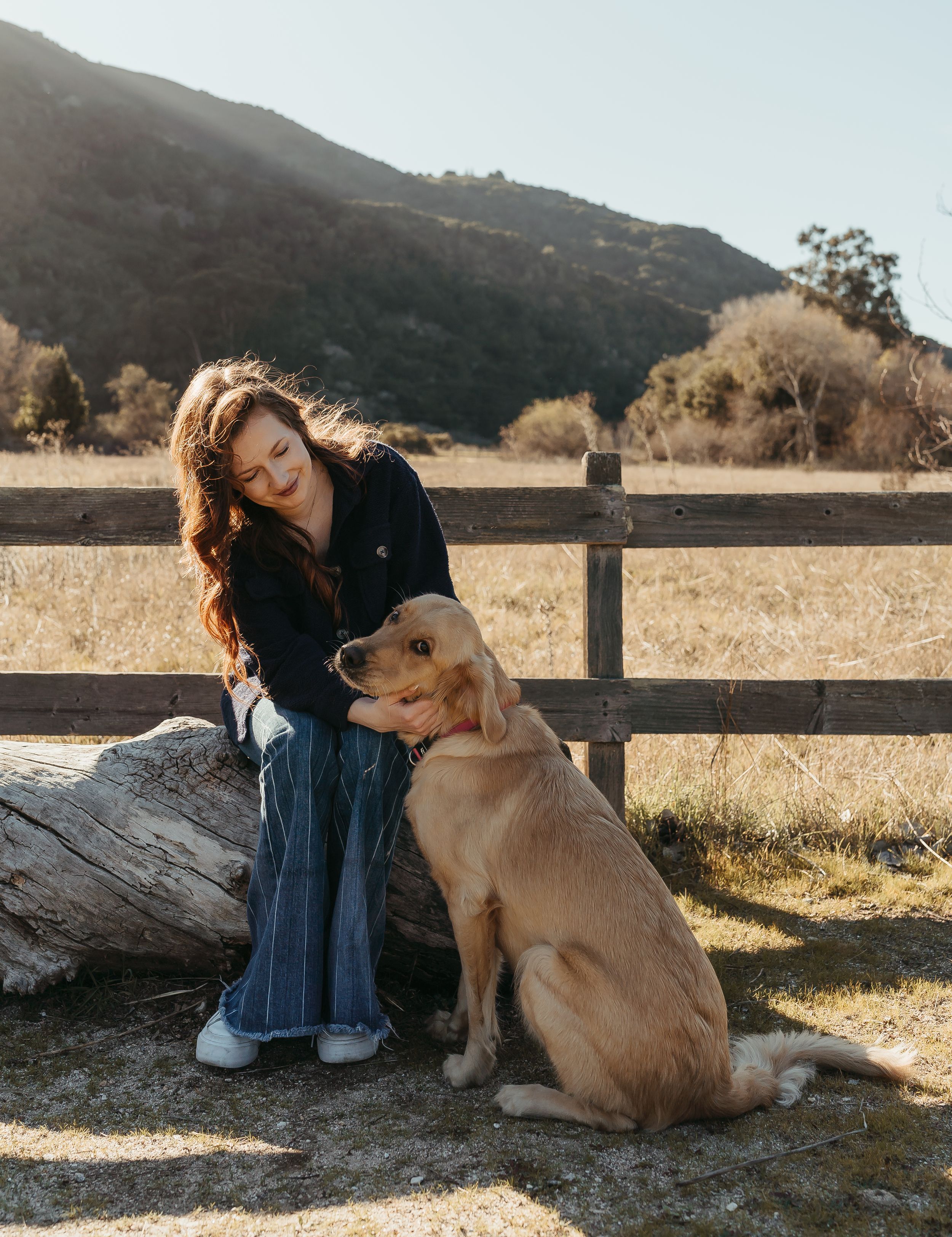 Woman in navy outfit sits on log petting golden retriever by wooden fence with mountain backdrop.