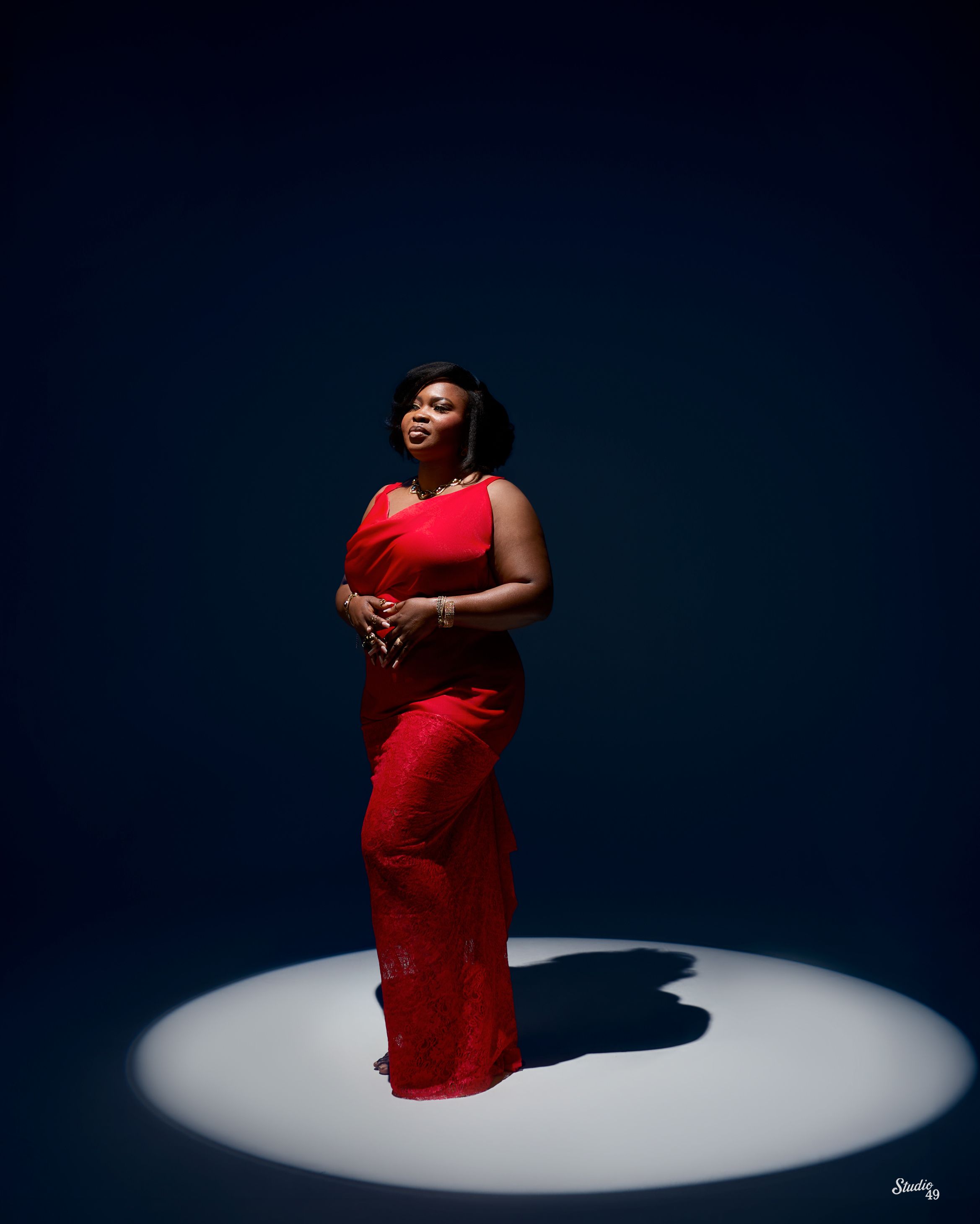 Confident woman in red gown standing in dramatic spotlight against dark blue background.
