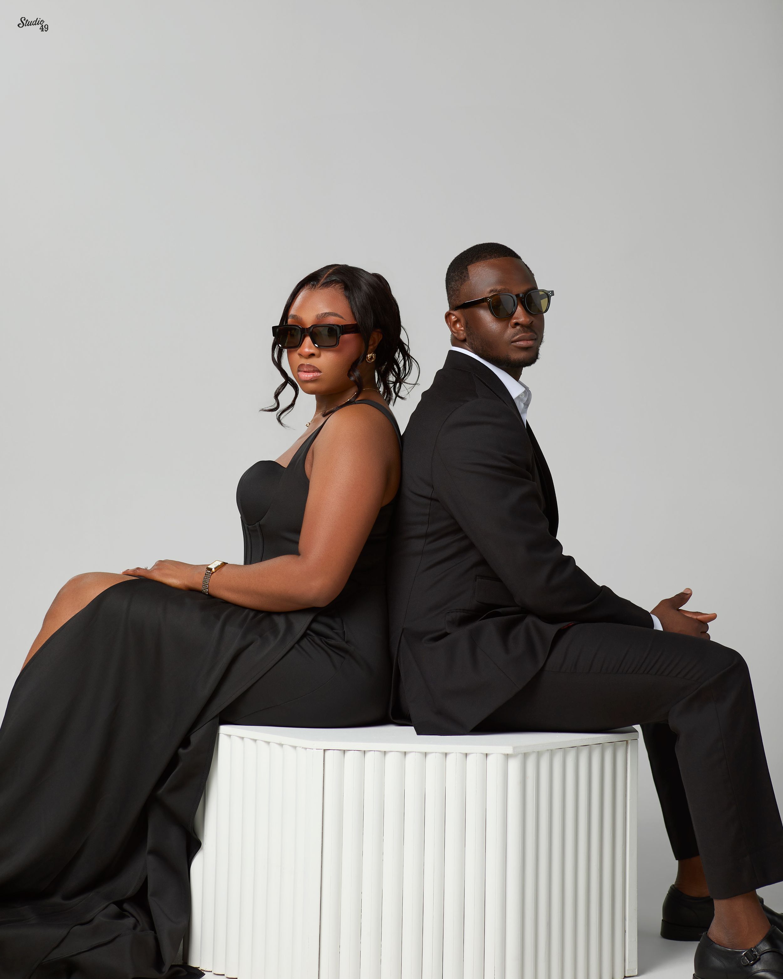 Stylish couple in matching black outfits posing back-to-back with sunglasses in studio.