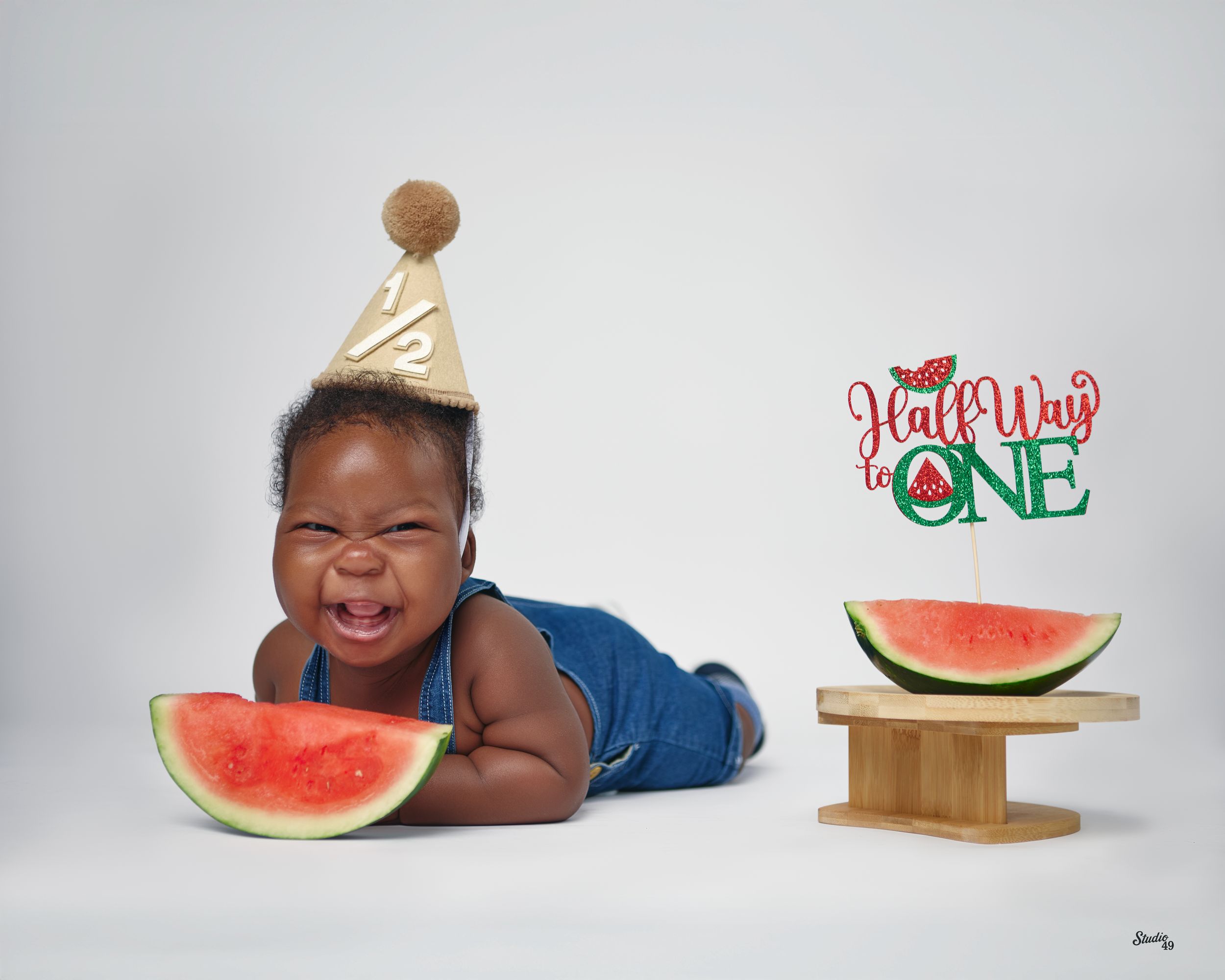 Adorable baby in denim overalls and party hat celebrating half birthday with watermelon slices.