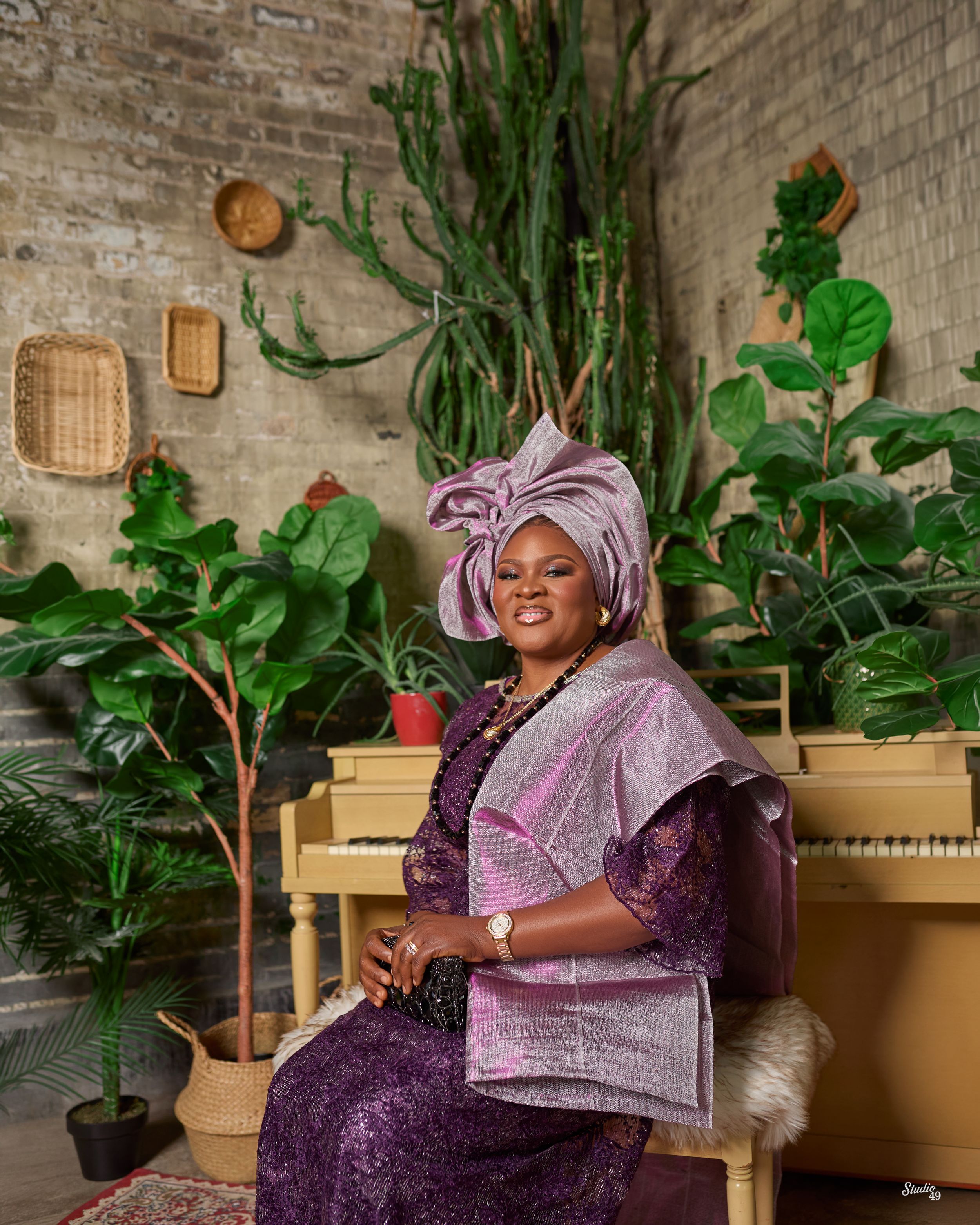 Elegant woman in purple lace outfit and pink gele headwrap seated among lush green plants.