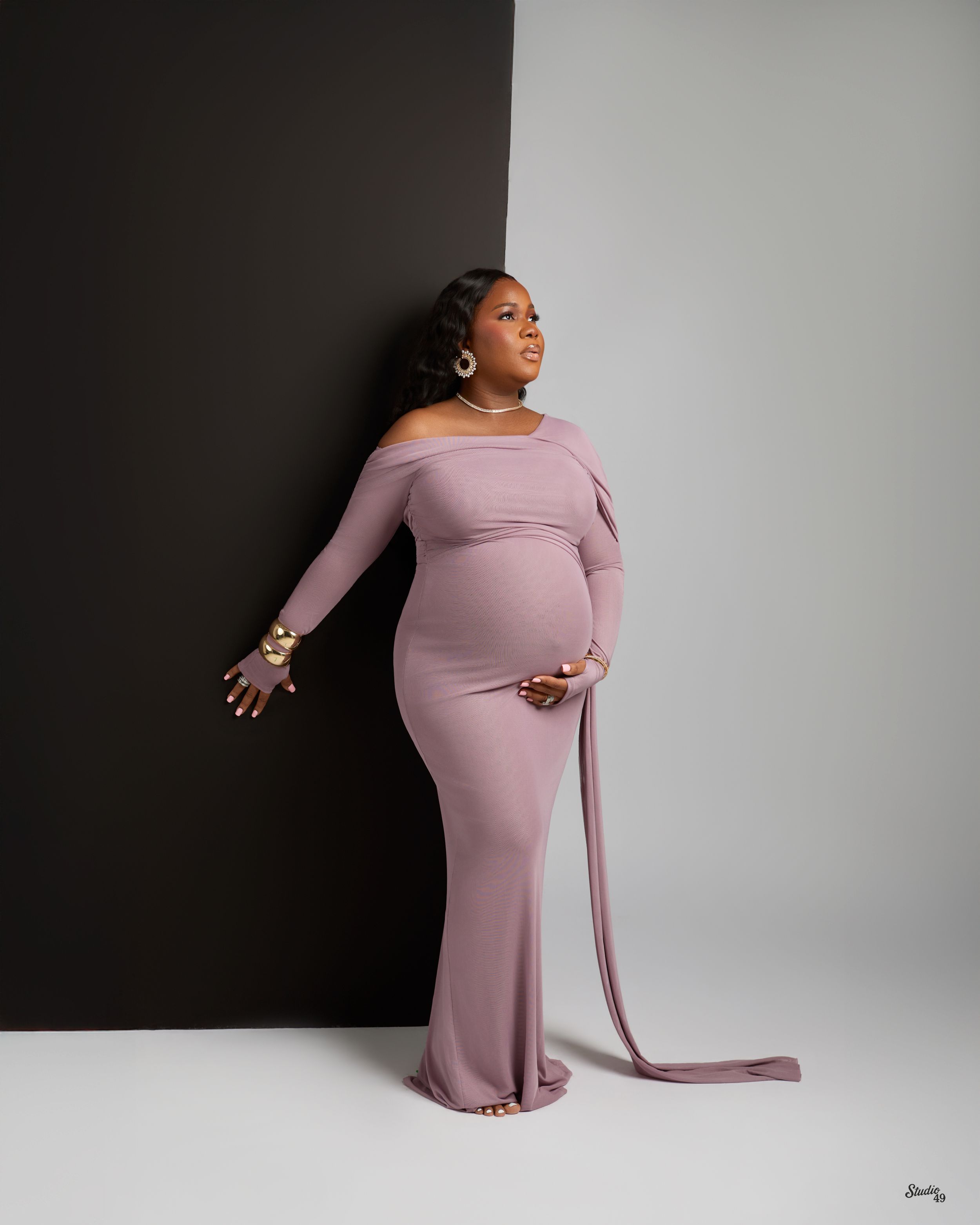 Elegant pregnant woman in mauve off-shoulder maternity gown posing in black and white studio.