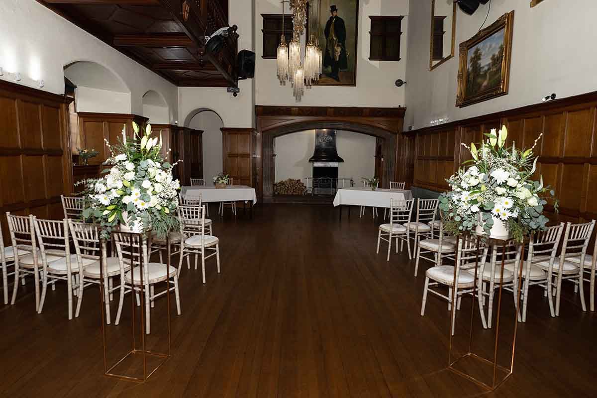 Elegant wedding ceremony setup in a historic wood-paneled hall with white floral arrangements and chiavari chairs at Dalston Hall